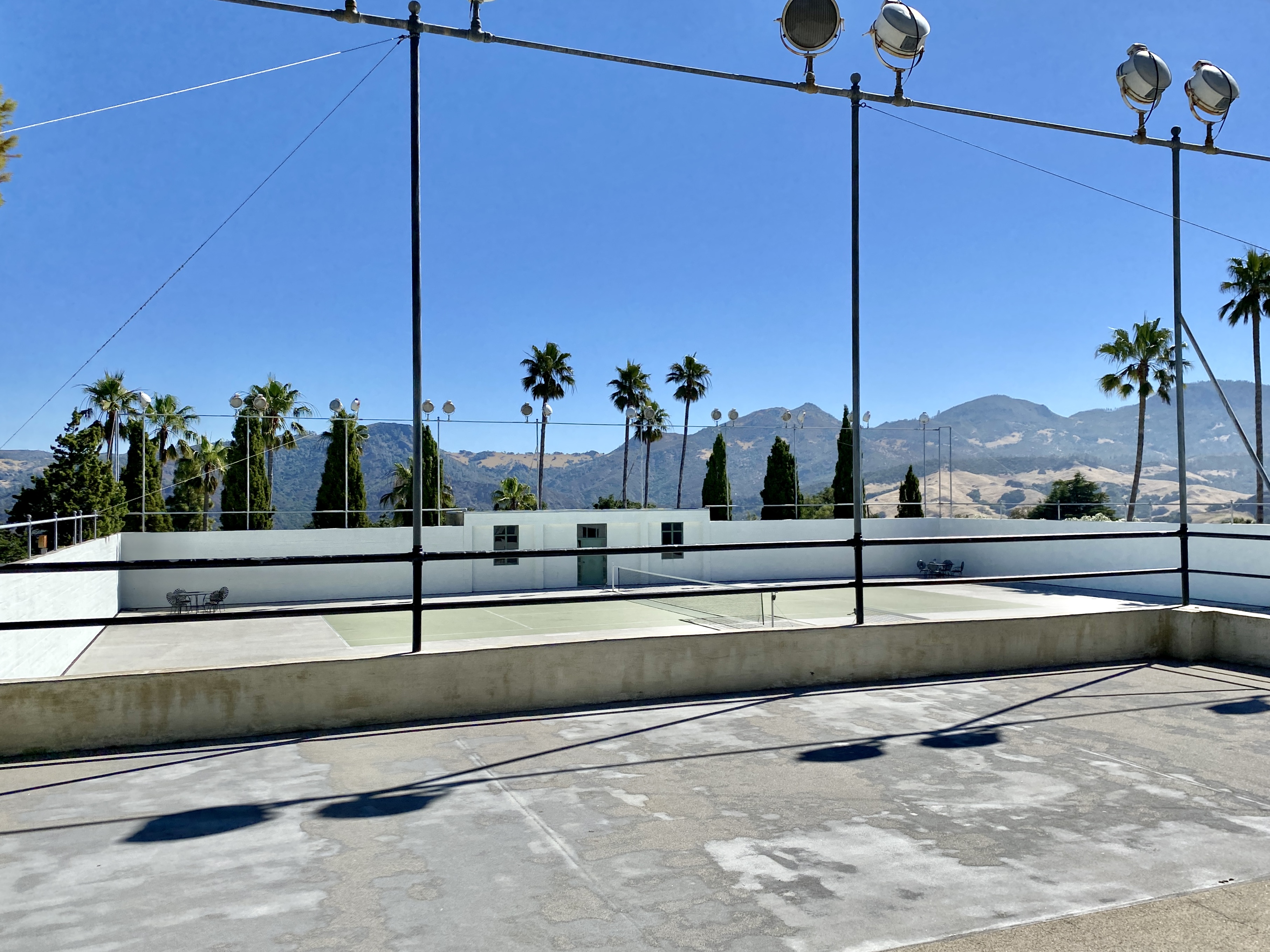 Outdoor tennis courts with tall light poles, palm trees, and mountains in the background.