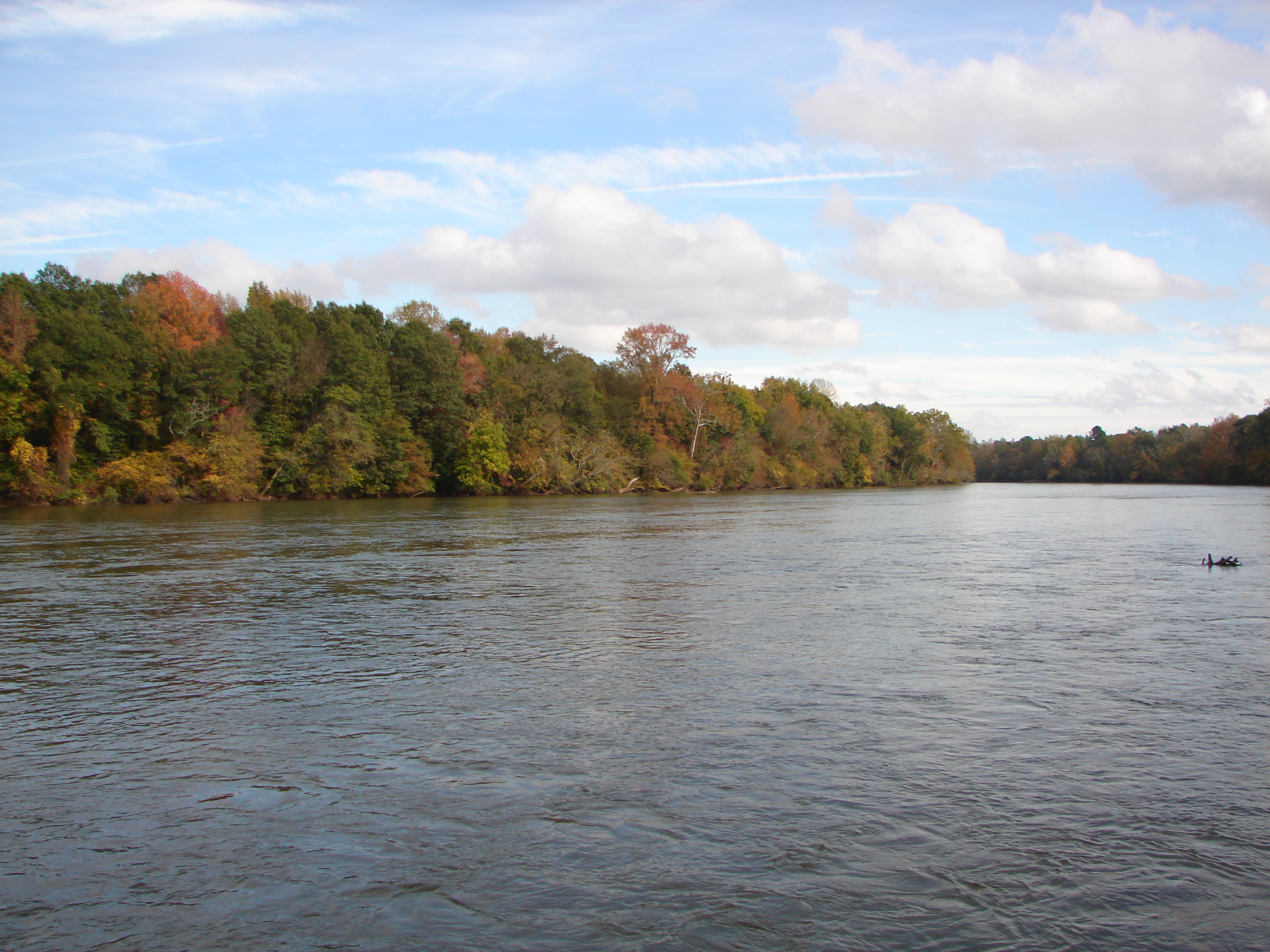 Wide river with calm water and tree-lined banks showing autumn foliage under a partly cloudy sky.