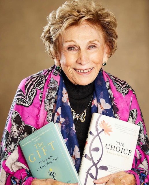 An elderly woman in a colorful floral jacket smiles while holding two hardcover books.