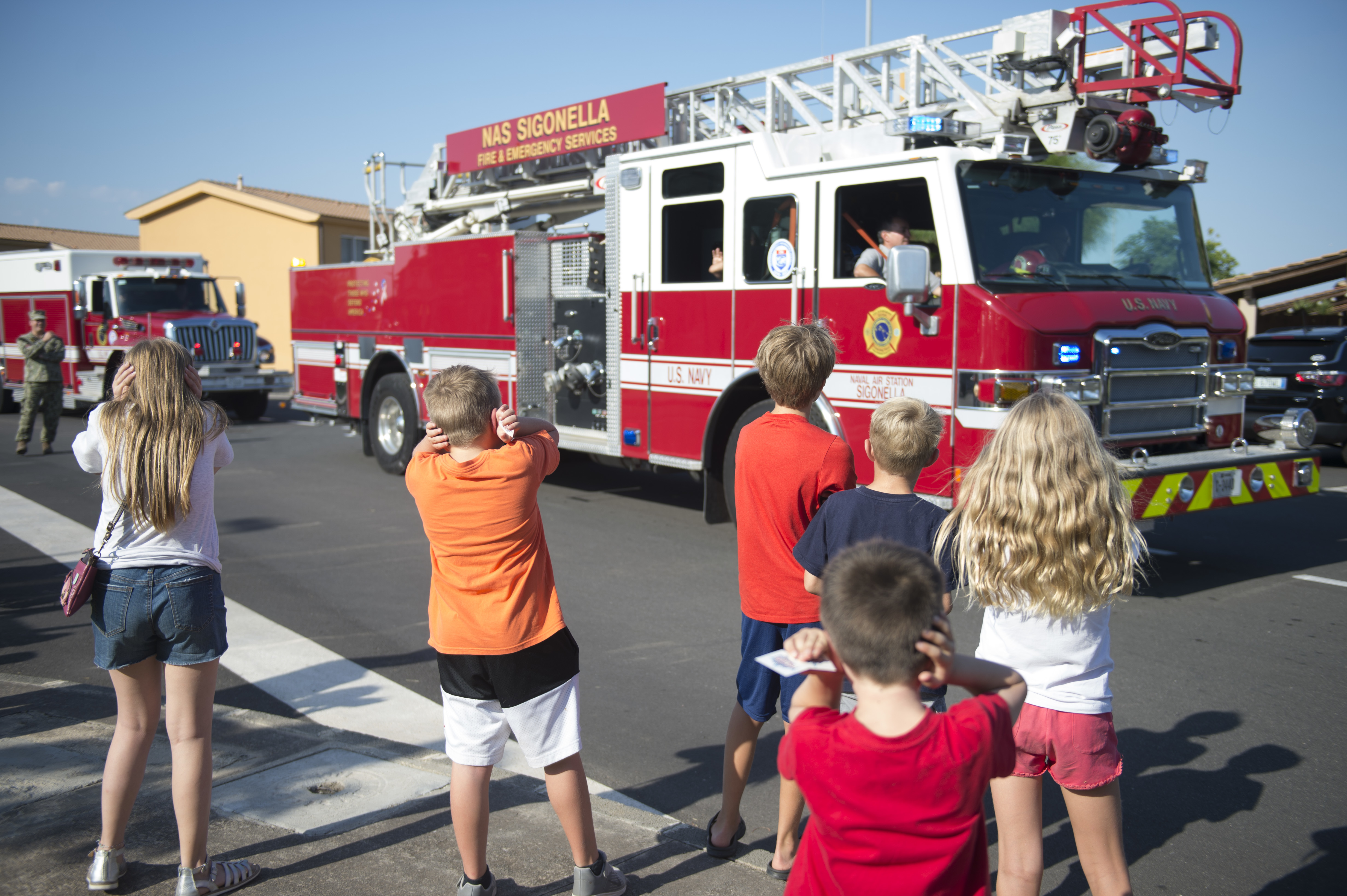 Children stand near a large red fire truck in a parking lot at an outdoor event.