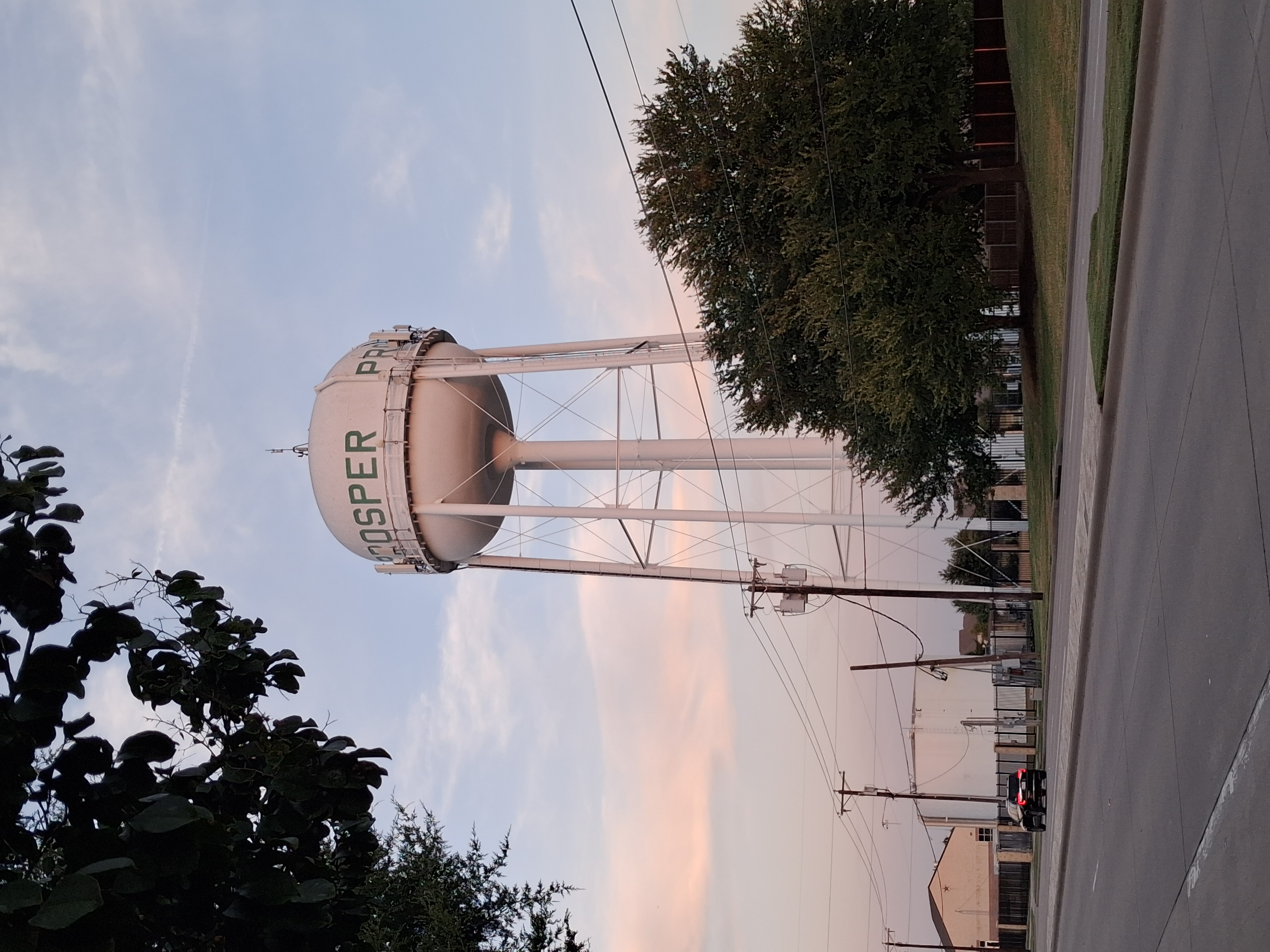 White spherical water tower on a steel support structure above trees and power lines at dusk.