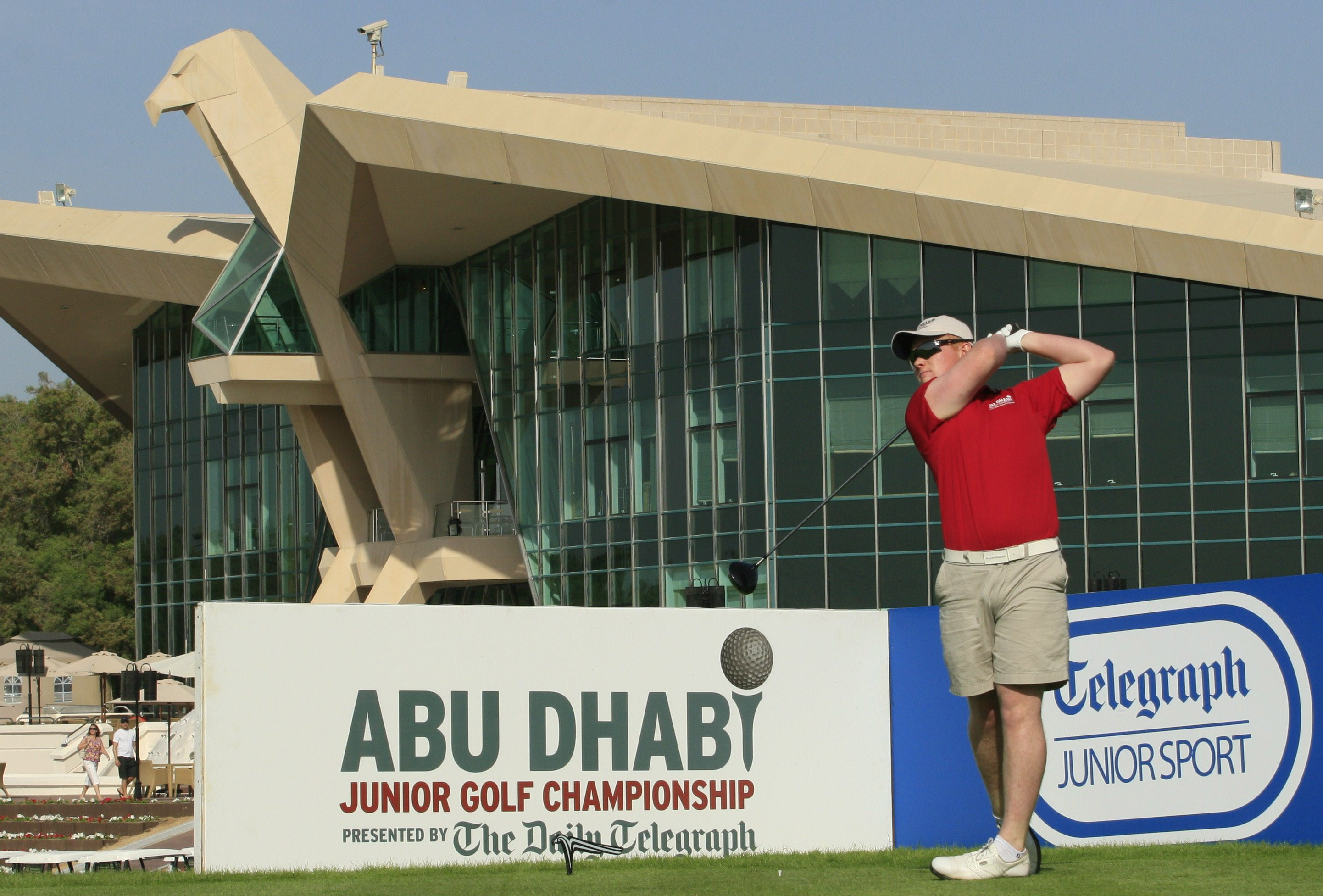 A young golfer in a red shirt completes a tee shot at a golf championship.