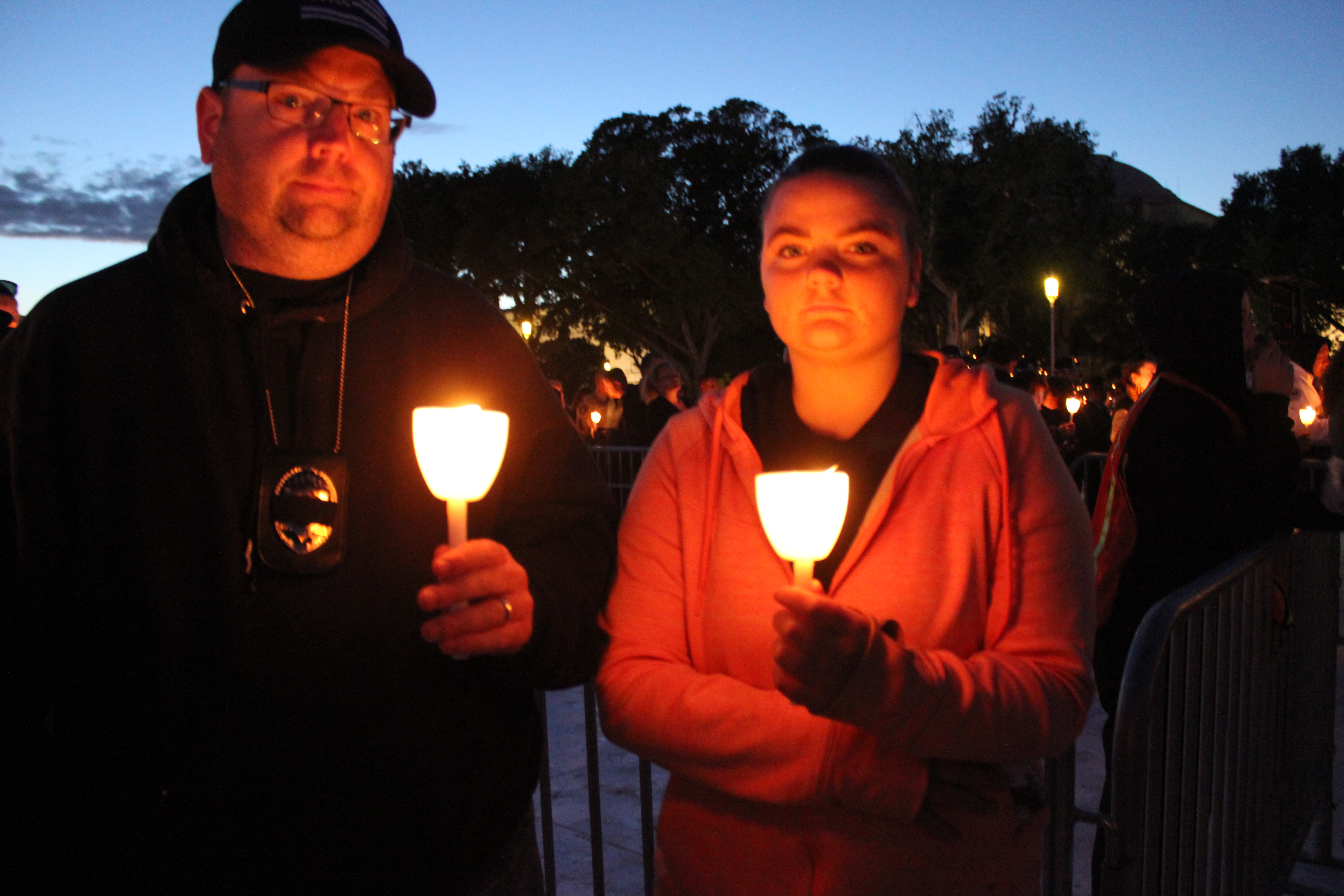 Two people holding lit candles at an outdoor candlelight vigil at dusk.