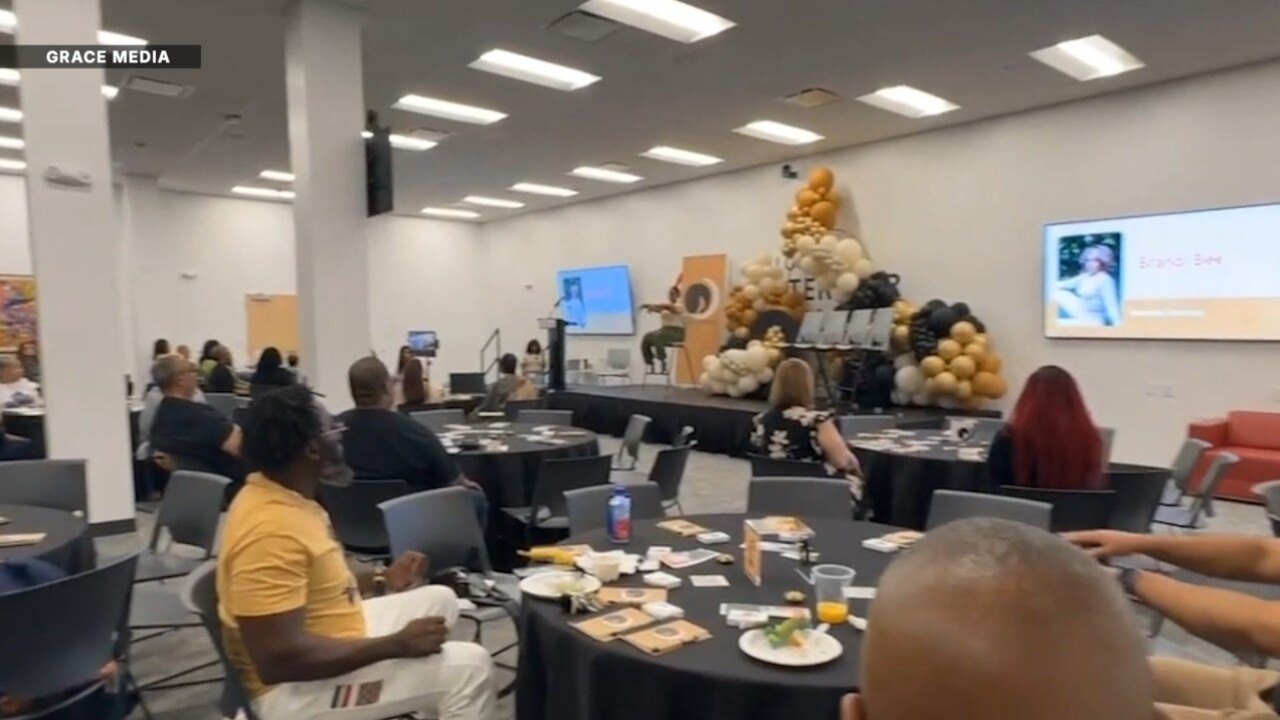 Conference room with attendees seated at round tables facing a speaker at a podium with gold, black, and white balloon decorations.