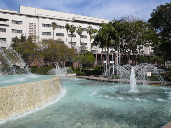 Tiered fountain with cascading water jets in front of a tall government building and palm trees.