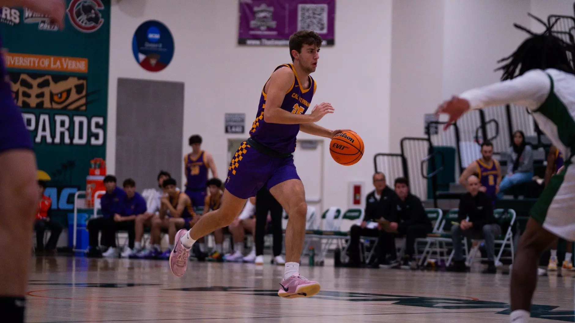 A basketball player in a purple and gold jersey dribbles on an indoor court.