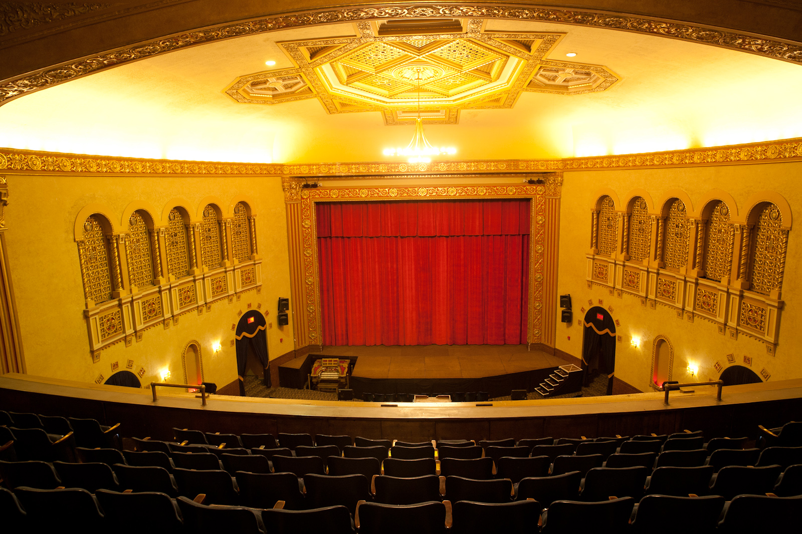 Theater interior viewed from the balcony, with rows of seats, red curtain stage, and ornate gold ceiling.