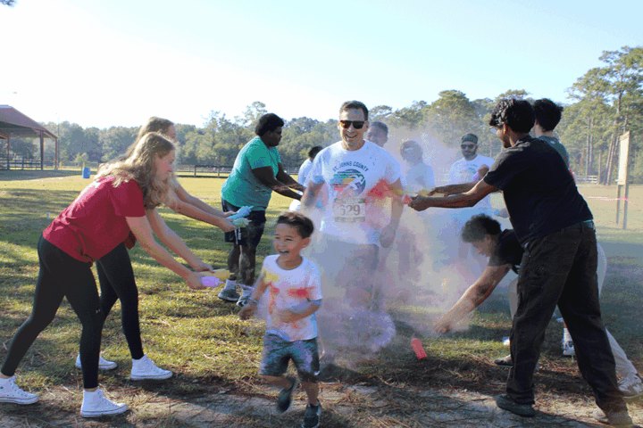 A child and adults run through a colorful powder cloud at an outdoor race.