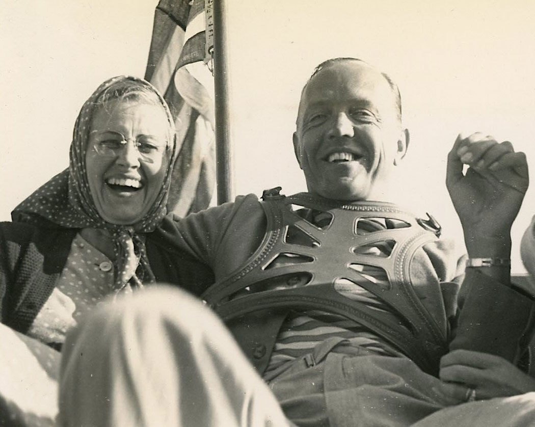 Black and white photo of a man and woman laughing together on a boat with a flag behind them.