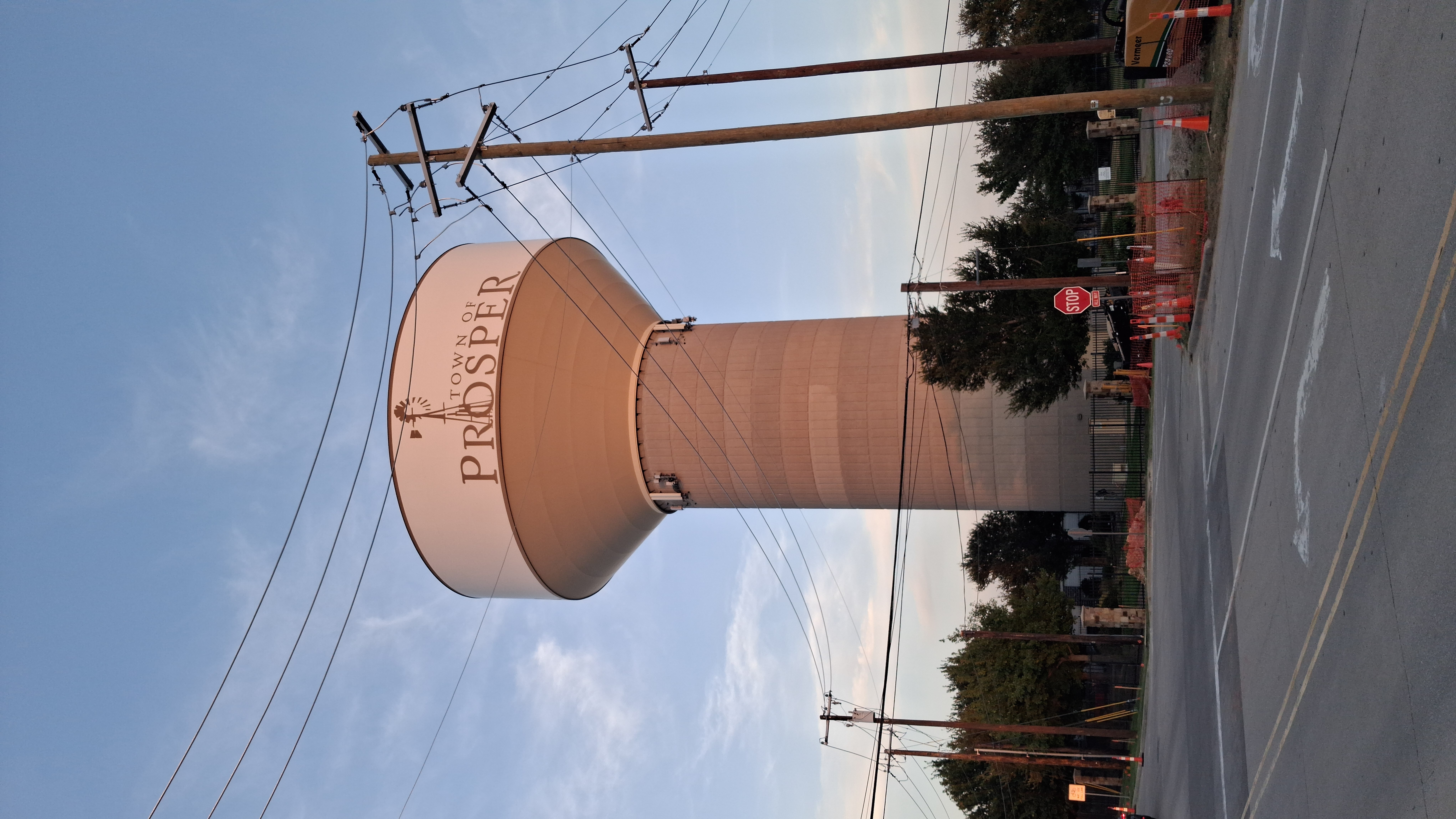 Tall municipal water tower labeled 'Town of Prosper' at dusk with utility poles and trees.