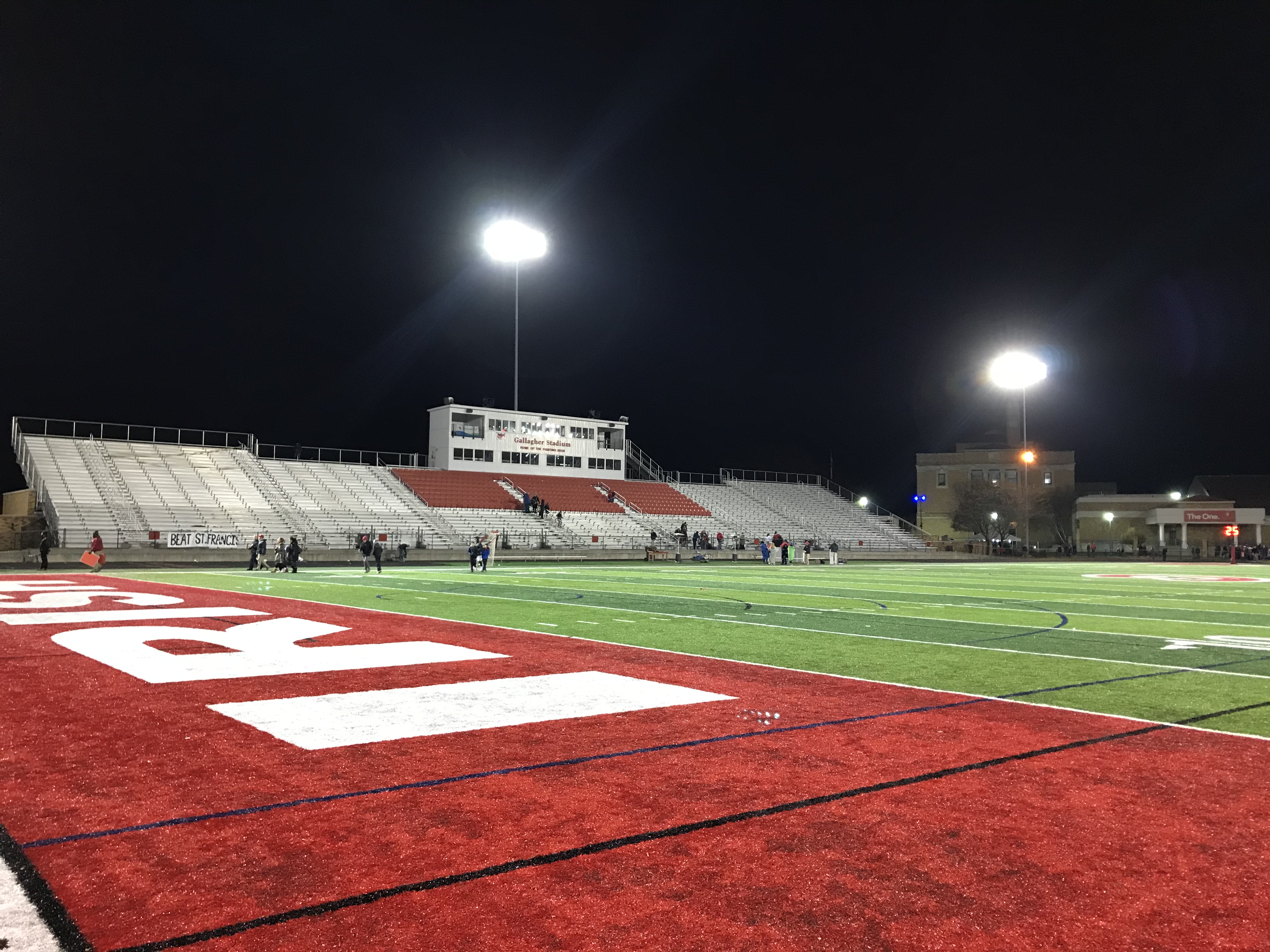High school football stadium lit up at night with red turf end zone and tall light towers.