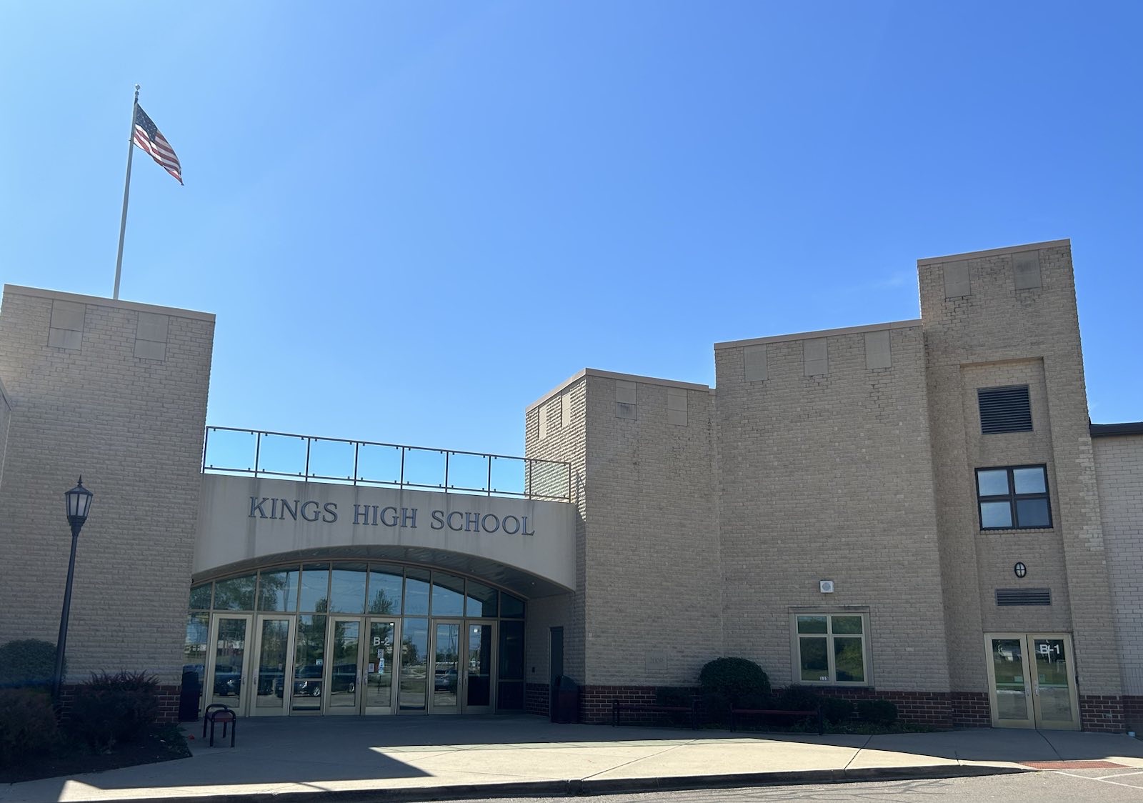 Tan brick school building with glass entrance doors and an American flag on a tall flagpole.