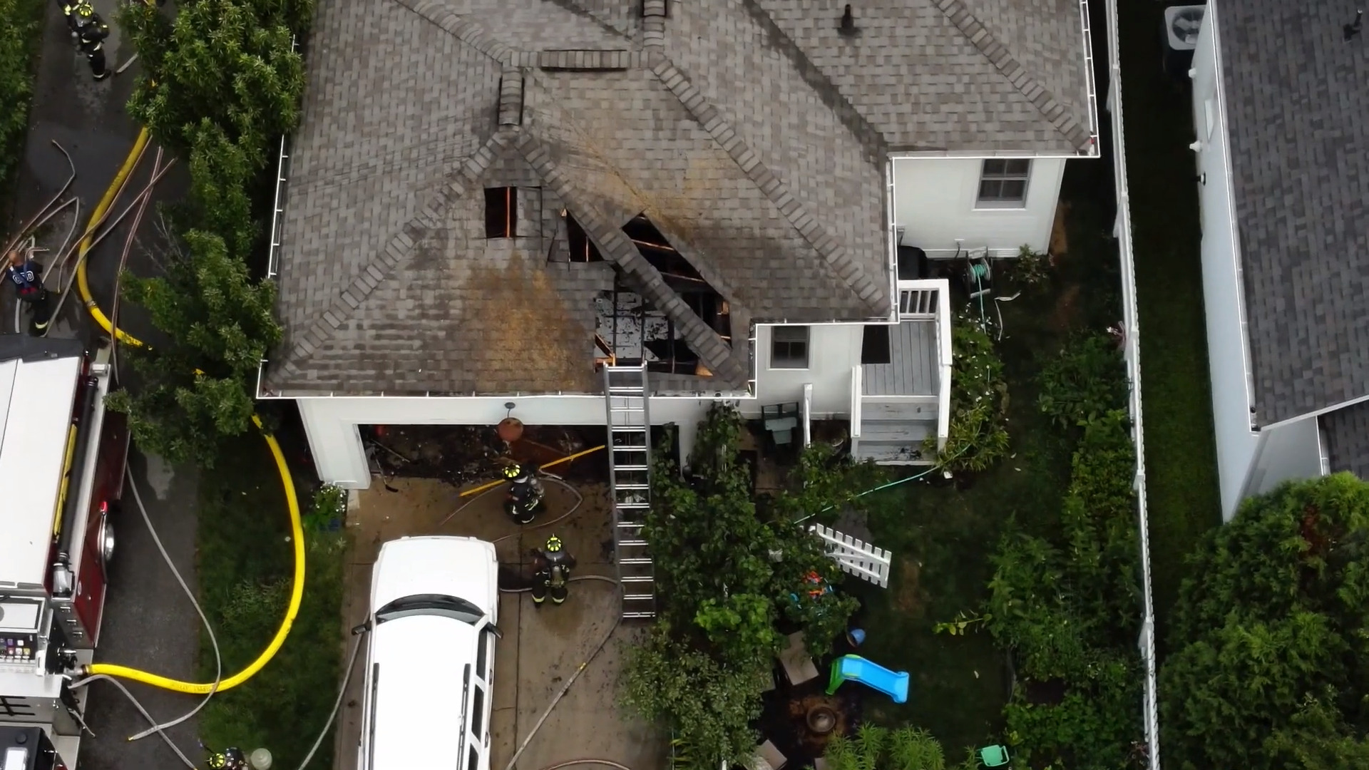 Aerial view of a house with a charred, fire-damaged roof and firefighters on scene.