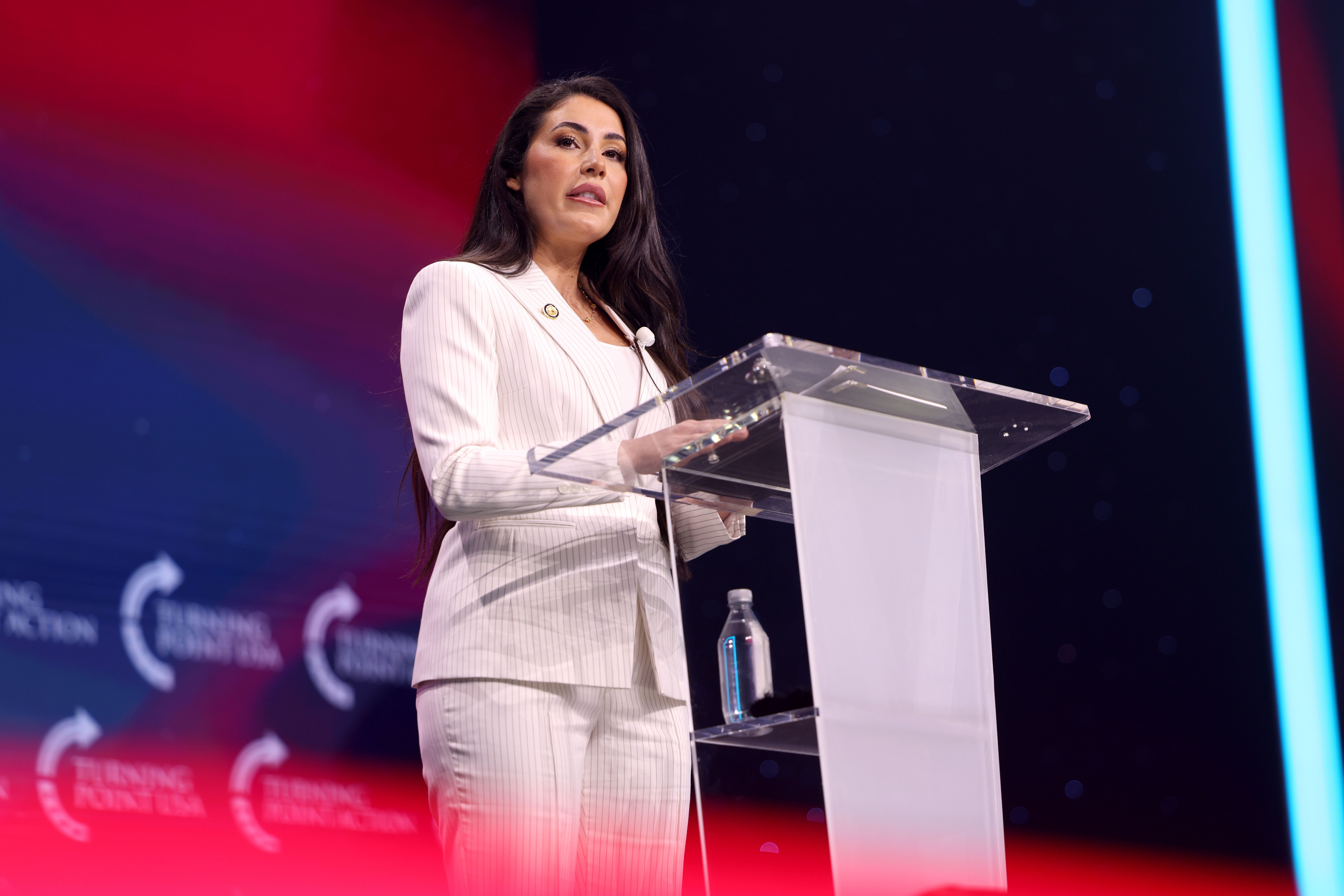 Woman in white pinstripe suit speaking at a clear podium before a red and blue lit backdrop.