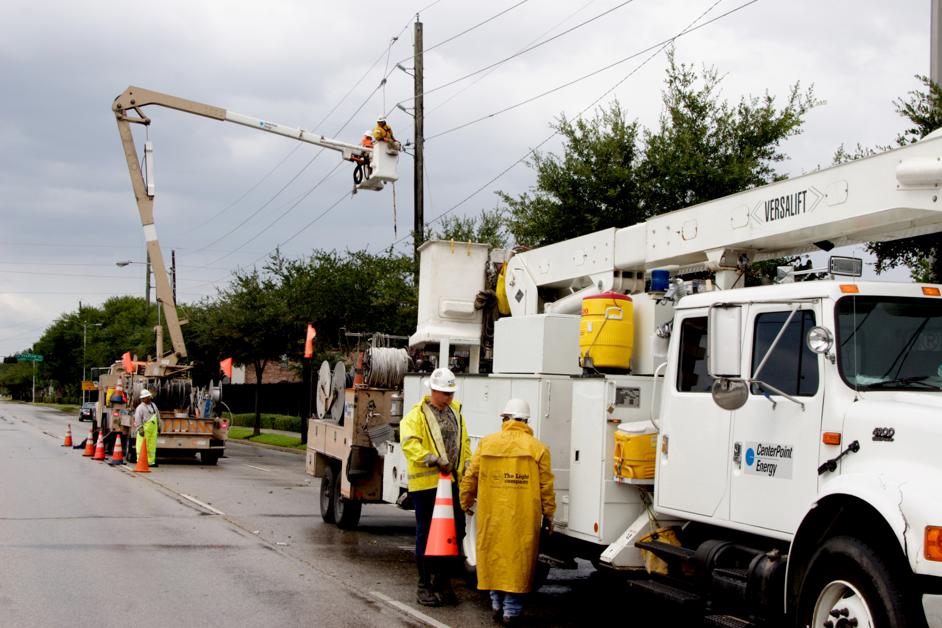 Utility workers in yellow rain gear repair overhead power lines from a bucket truck.