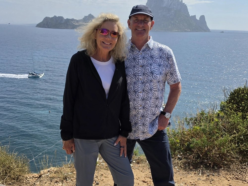 A smiling couple stands together on a rocky coastal overlook with ocean water behind them.