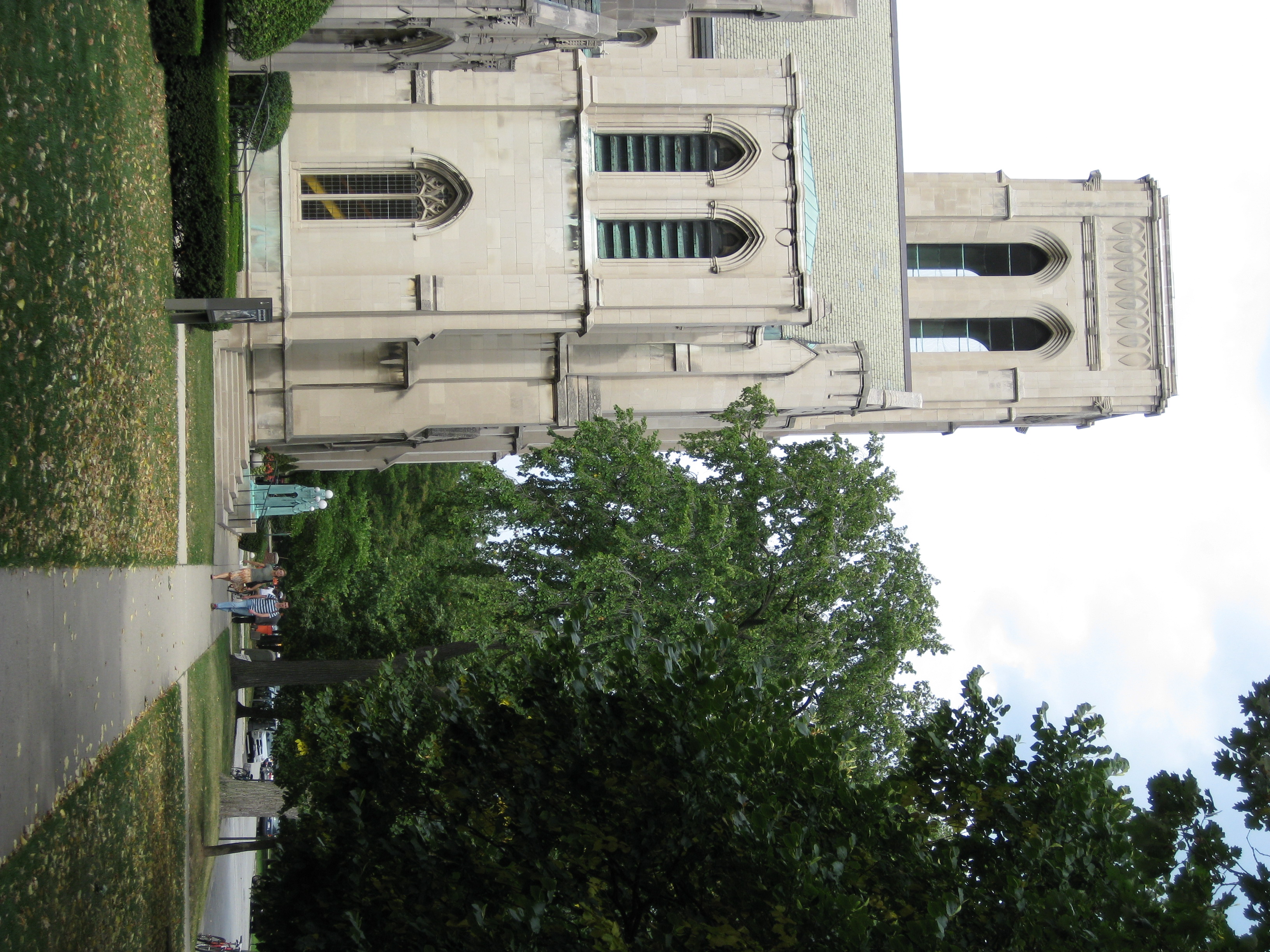 Gothic Revival stone church with a tall tower, arched windows, trees, and pedestrians on a walkway.