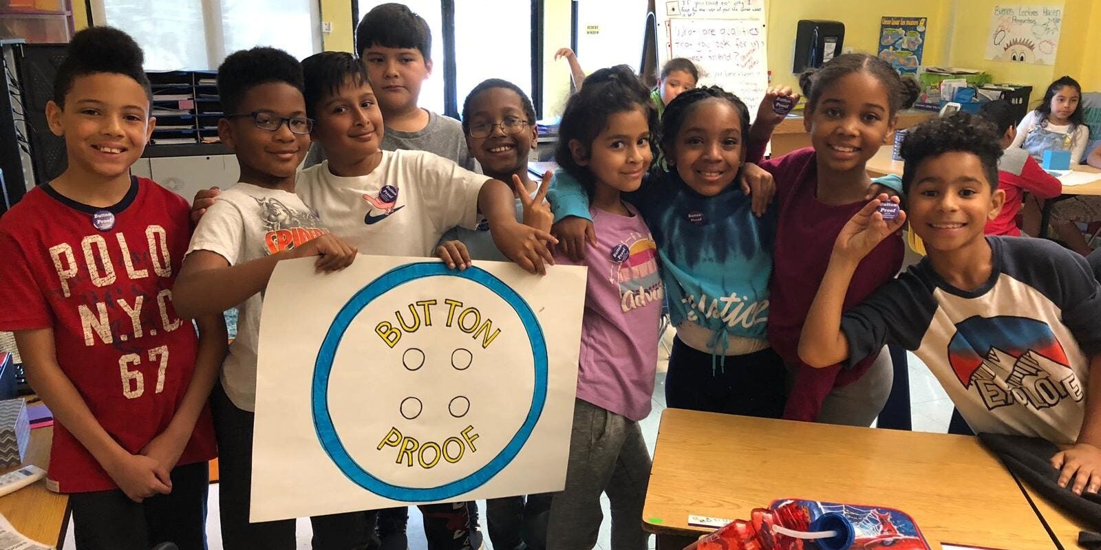 A group of children in a classroom hold a hand-drawn sign reading 'Button Proof.'