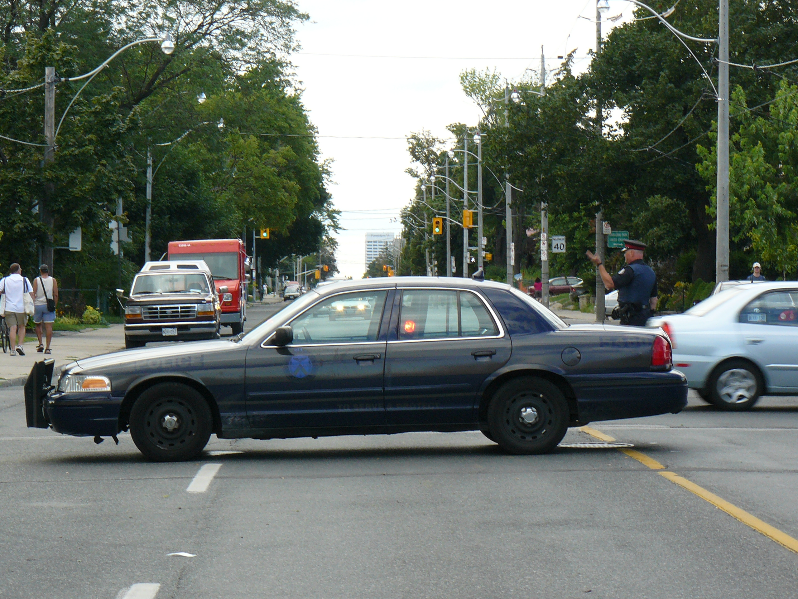 A damaged dark blue sedan at an intersection with a uniformed officer standing nearby.