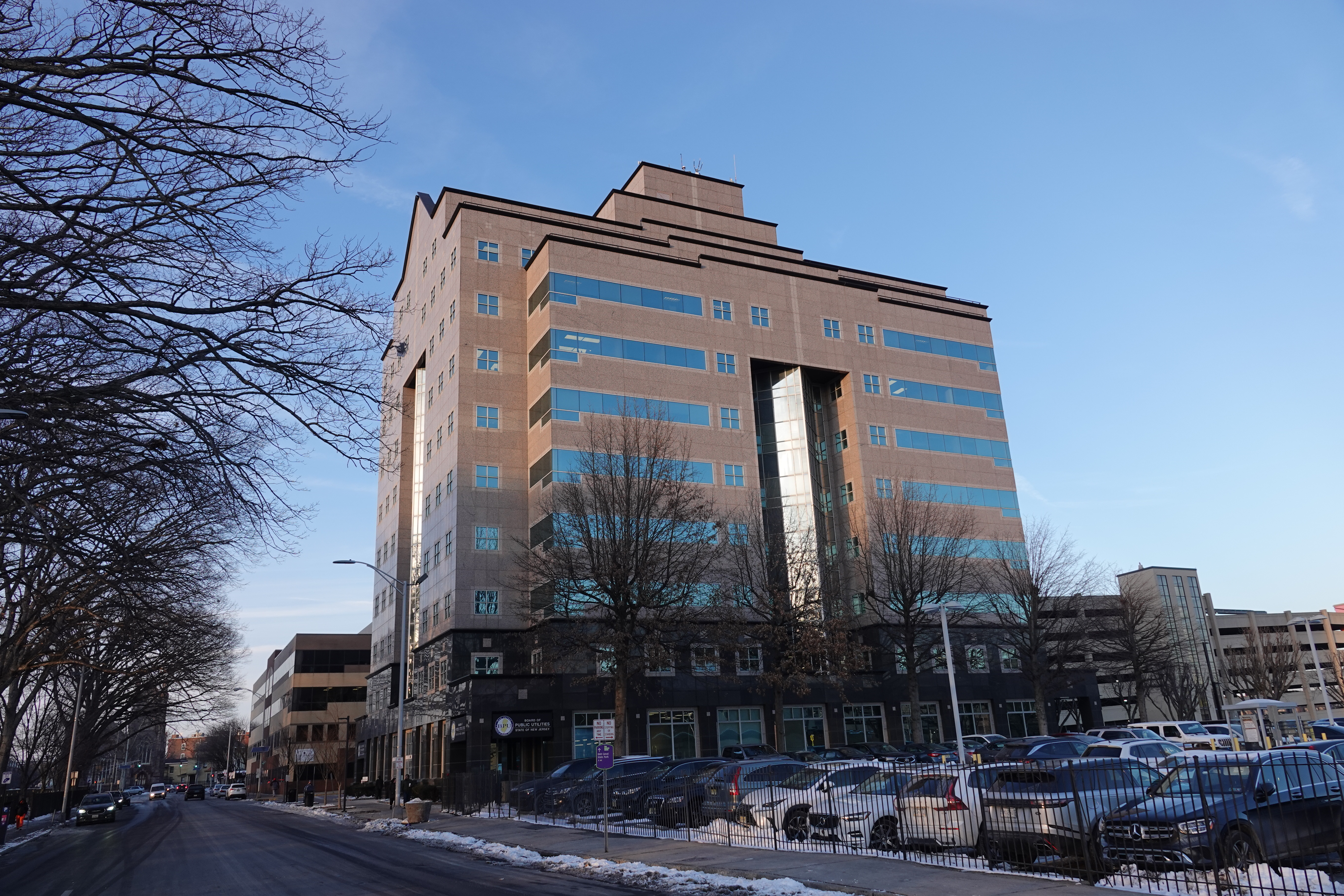 Tan brick and glass office building in winter, snow on the ground, bare trees in foreground.