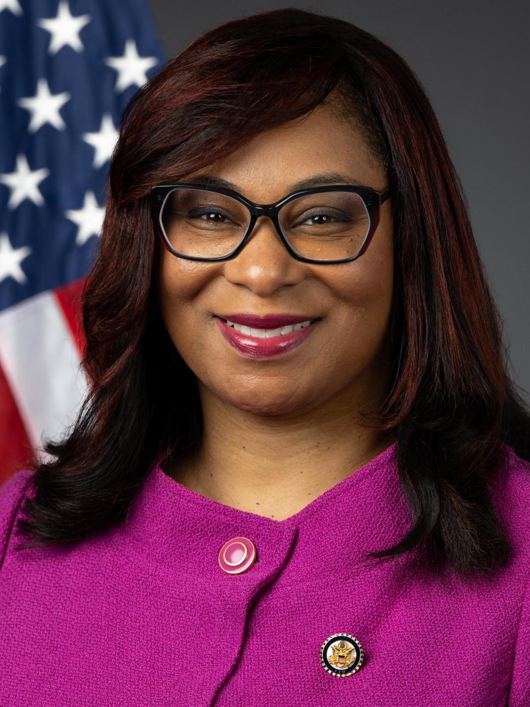 Woman in a magenta blazer and glasses posed before a U.S. flag.