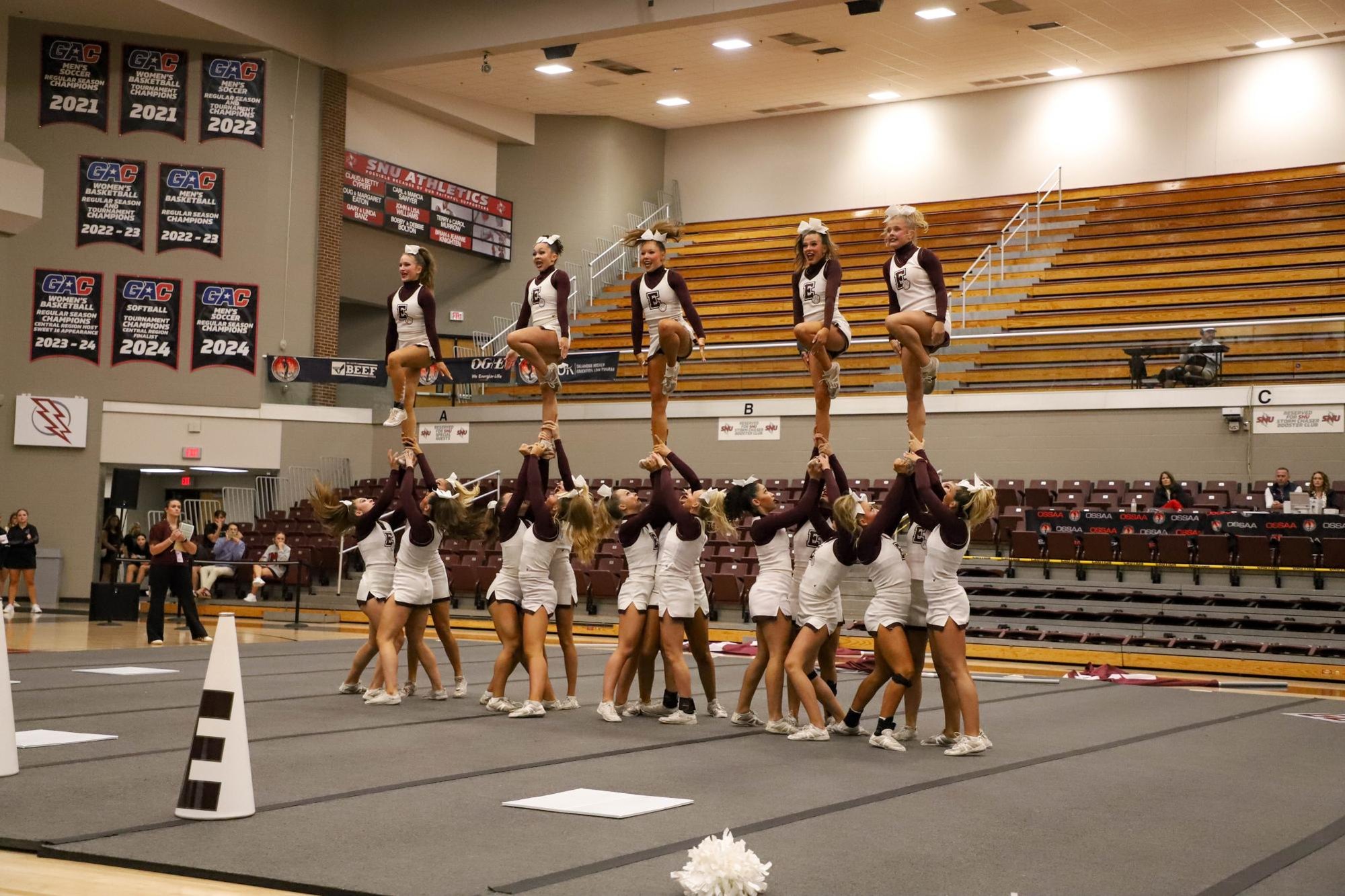 Cheerleaders in maroon and white uniforms form a multi-level pyramid stunt in a gymnasium.
