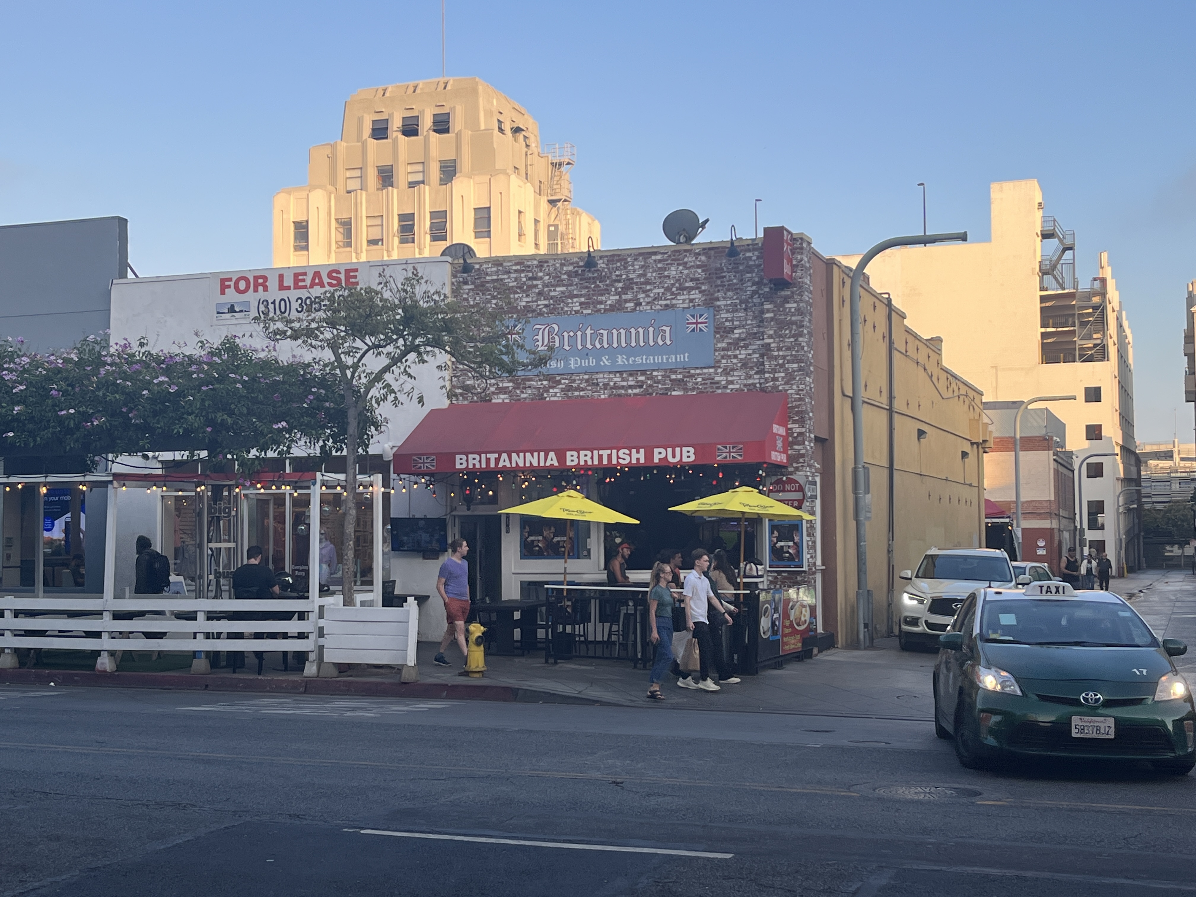 Exterior of Britannia British Pub with yellow umbrellas and a For Lease sign on a neighboring building.