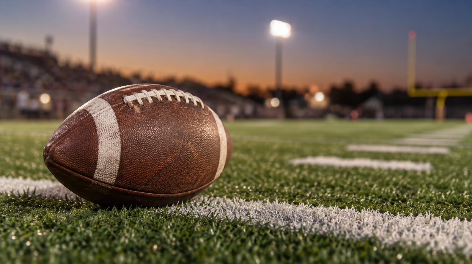 An American football on green artificial turf beside a white yard line