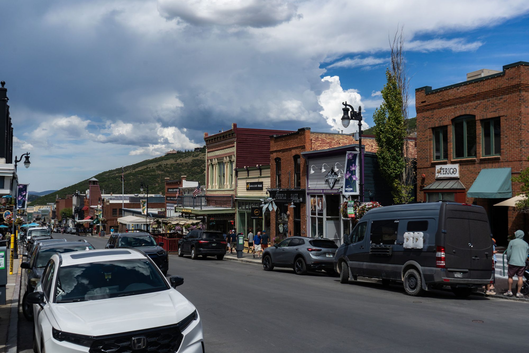 Busy small-town main street with brick storefronts, parked vehicles, and a tree-covered hillside.