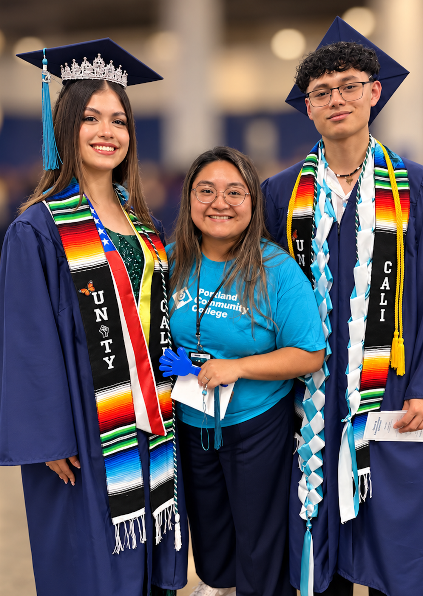 Two graduates in navy gowns and colorful stoles pose with a college staff member.