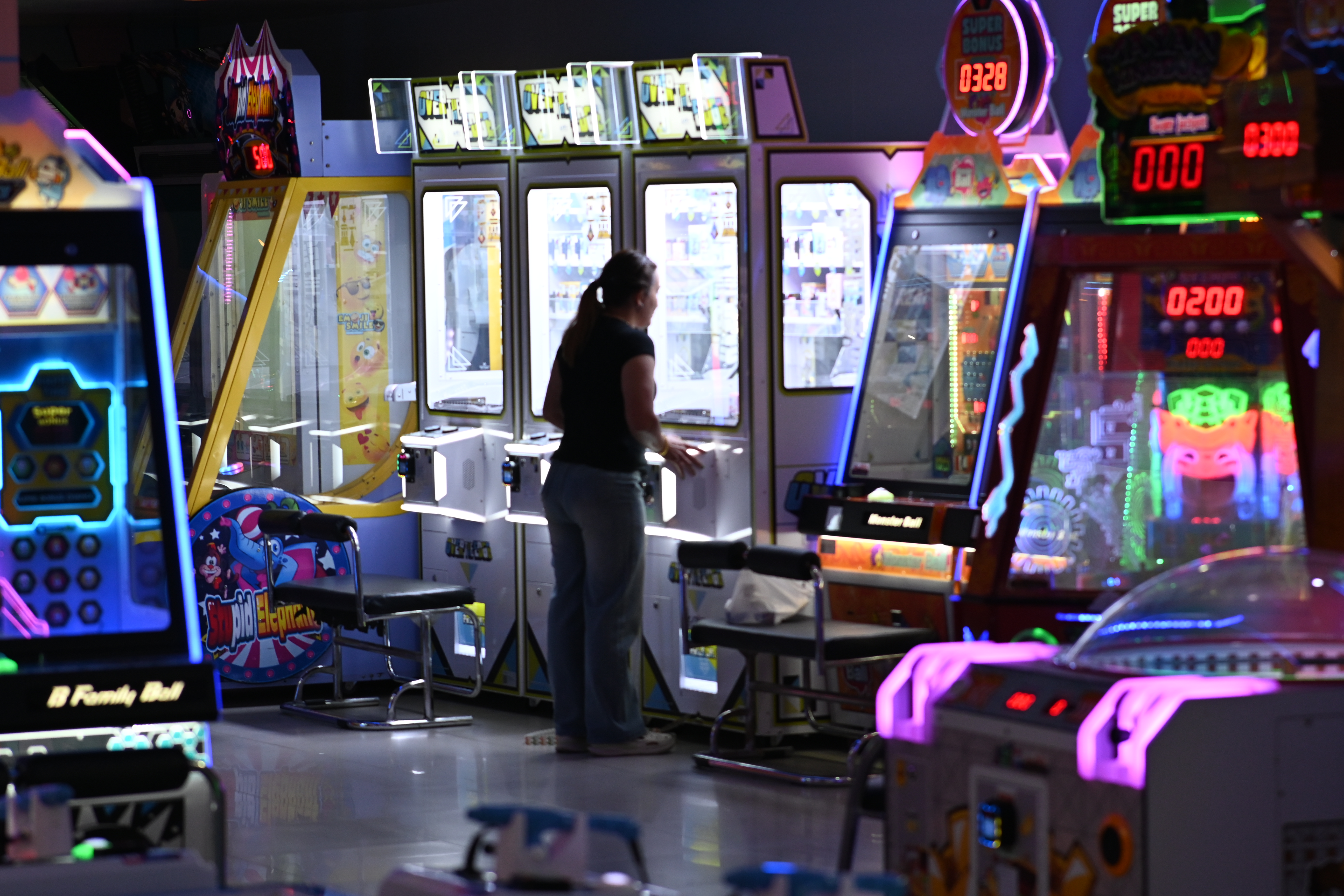 Person at a claw machine in a dark arcade lit by colorful neon and LED displays.