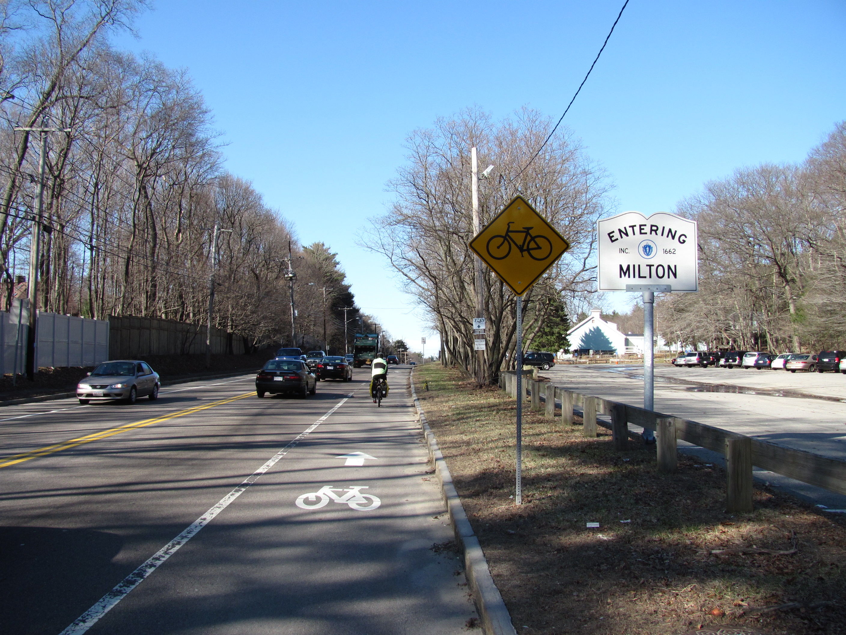 Road sign reading 'Entering Milton' on a multi-lane road lined with bare trees.