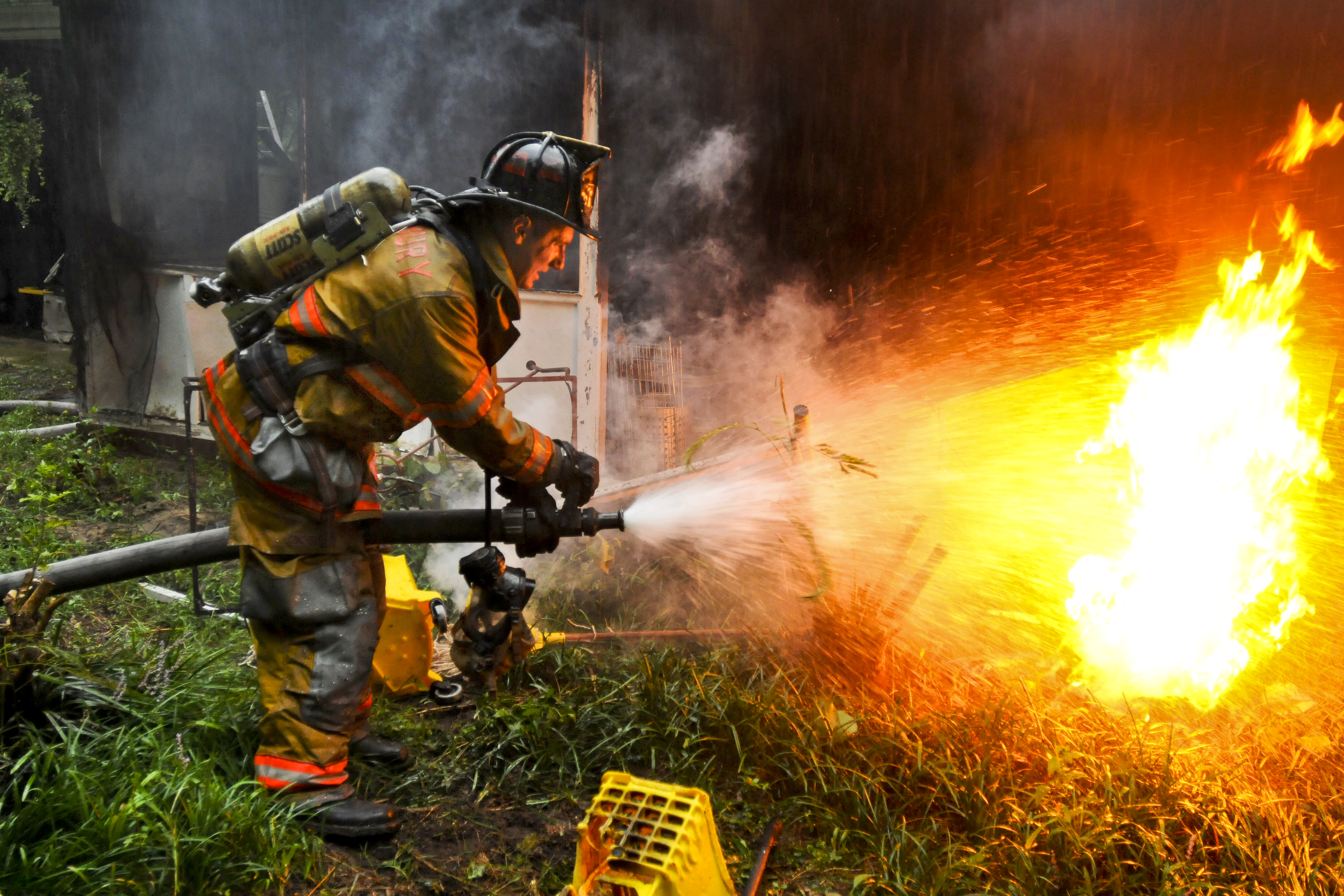 Firefighter in turnout gear crouches and aims a hose at large orange flames.