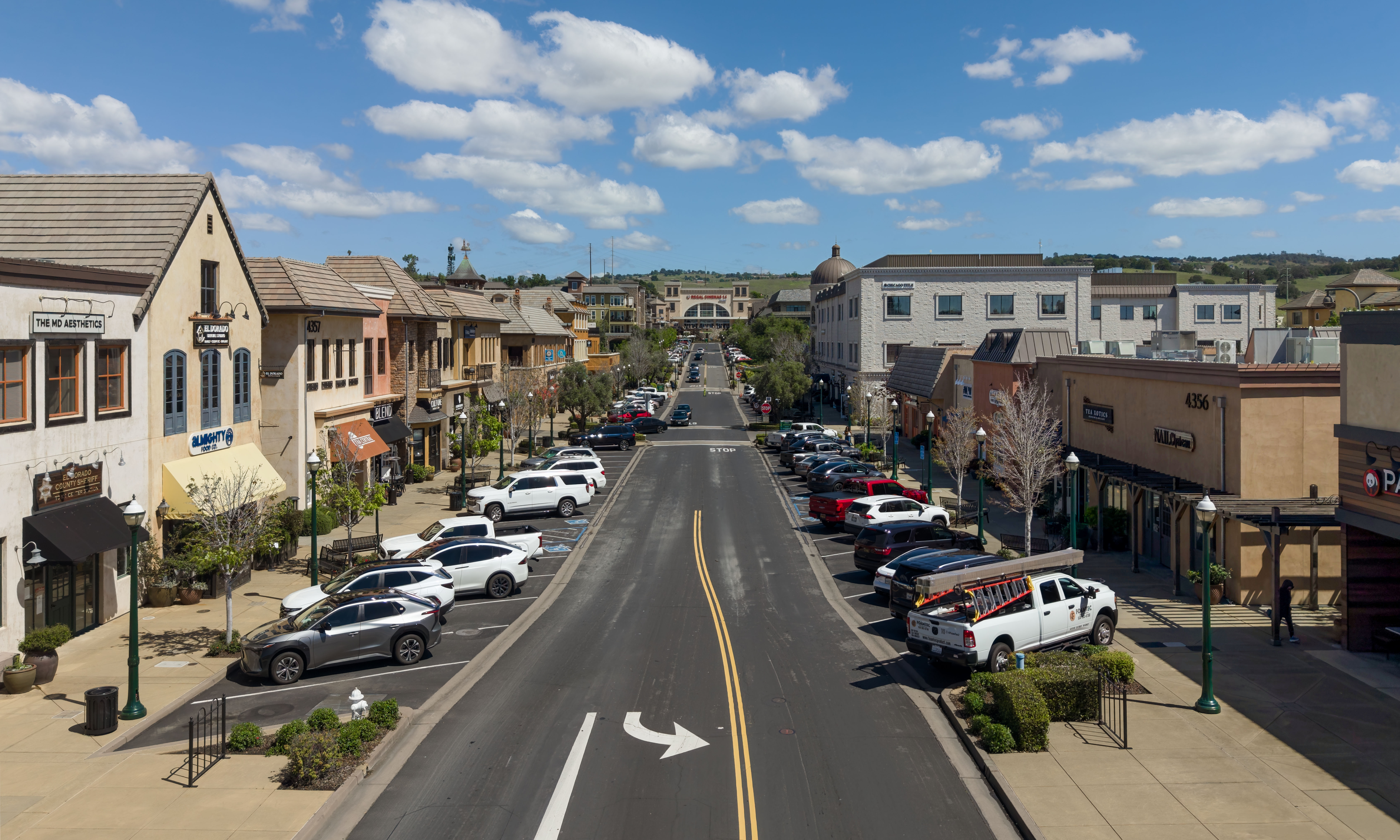 Wide commercial street with storefronts, parked cars, and rolling hills in the background.