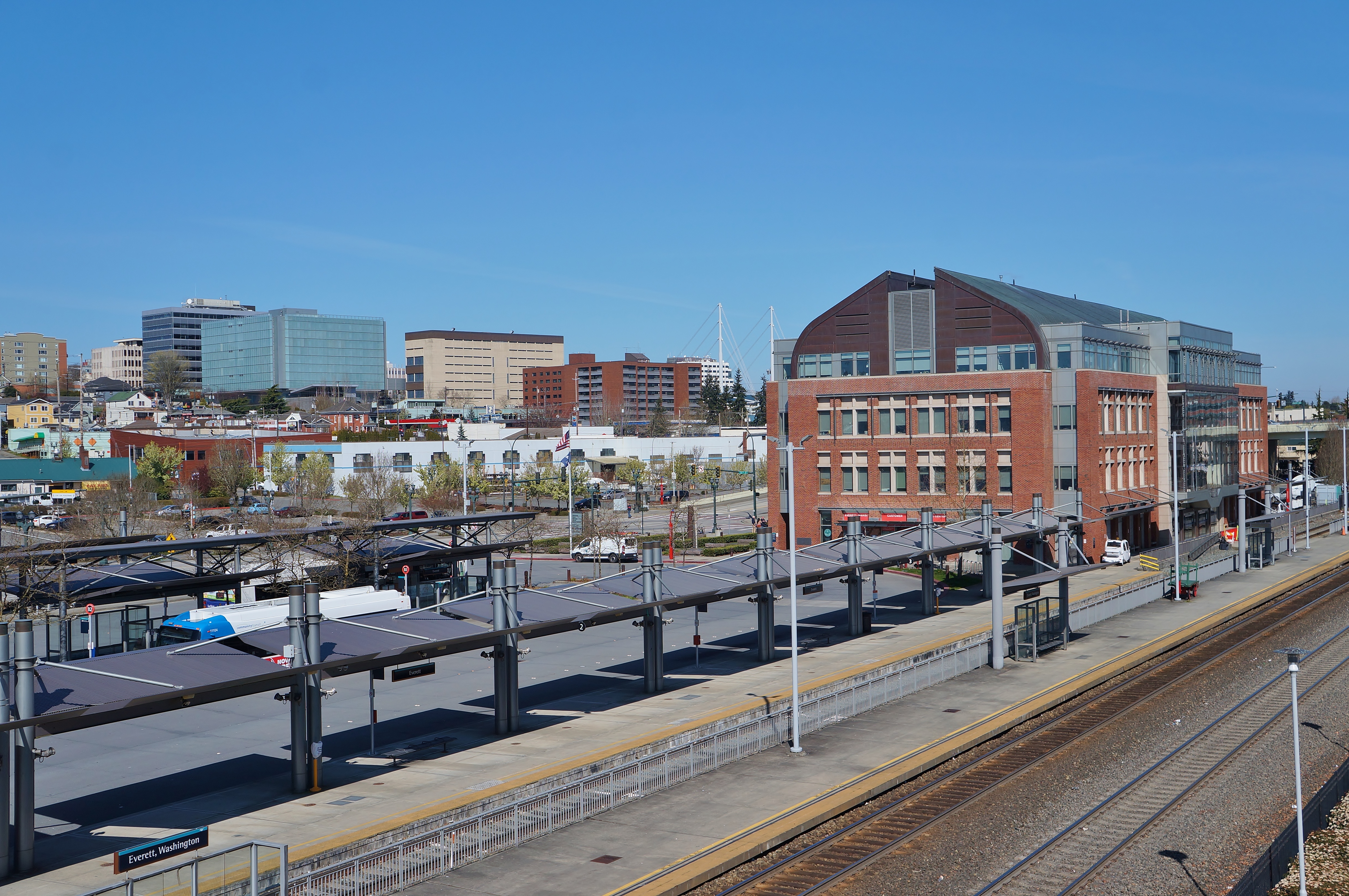 Train station platform and tracks in foreground with a brick station building and city skyline behind.