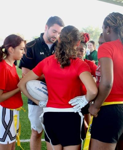 A coach in a black polo huddles with female athletes in red jerseys holding a football.