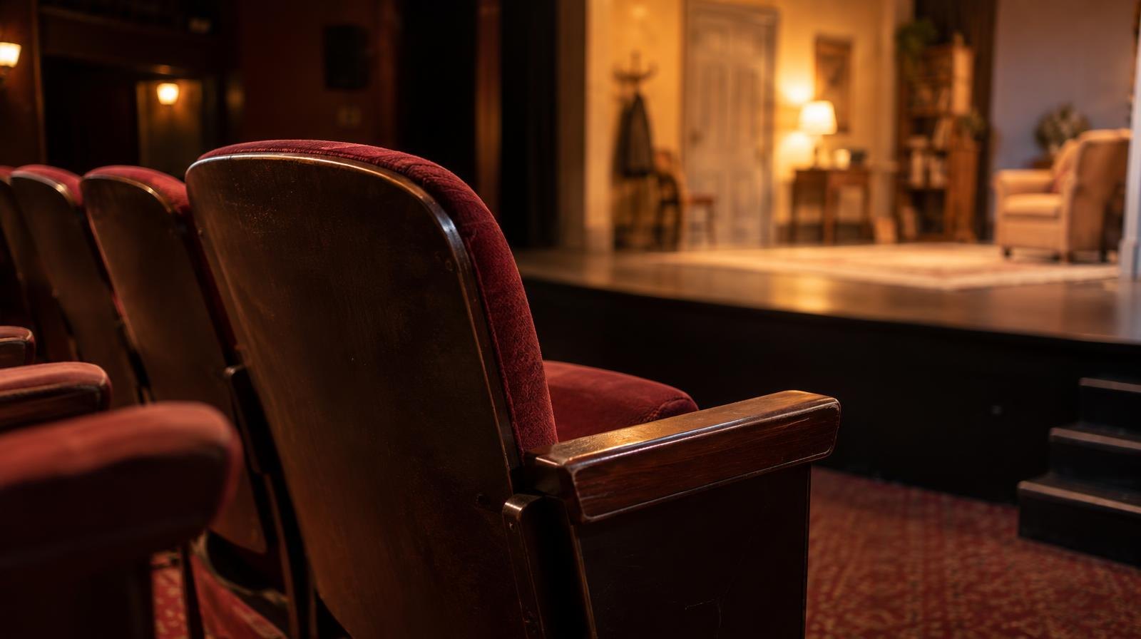 Empty theater seats facing a warmly lit stage in a small playhouse.
