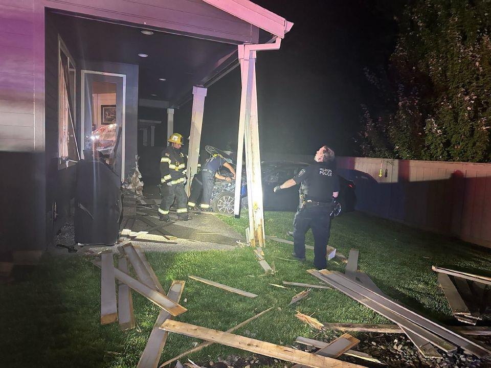Damaged home exterior at night with a police officer and two firefighters amid scattered debris.