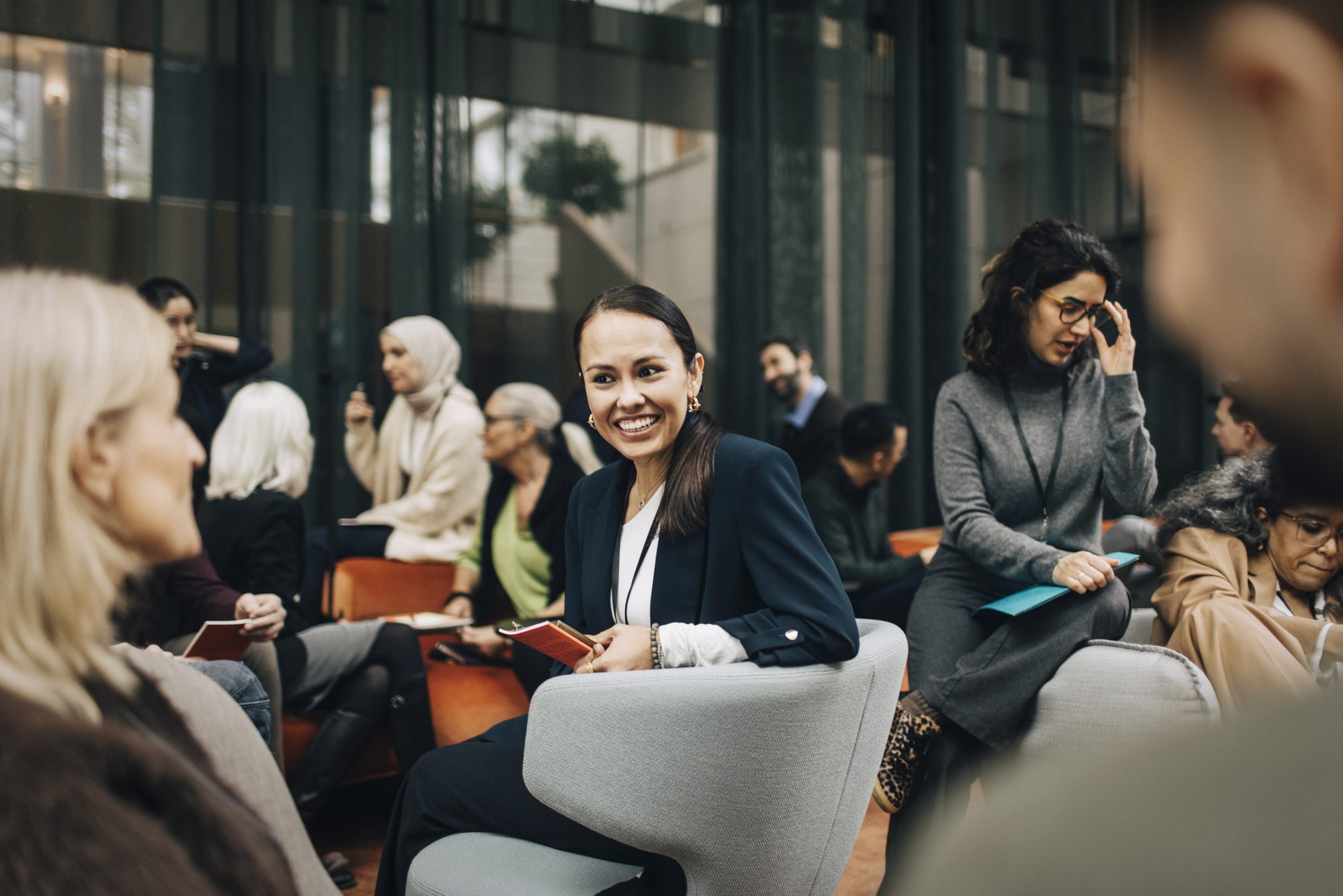 Smiling businesswoman discussing with colleagues while sitting on armchair during seminar in congress center