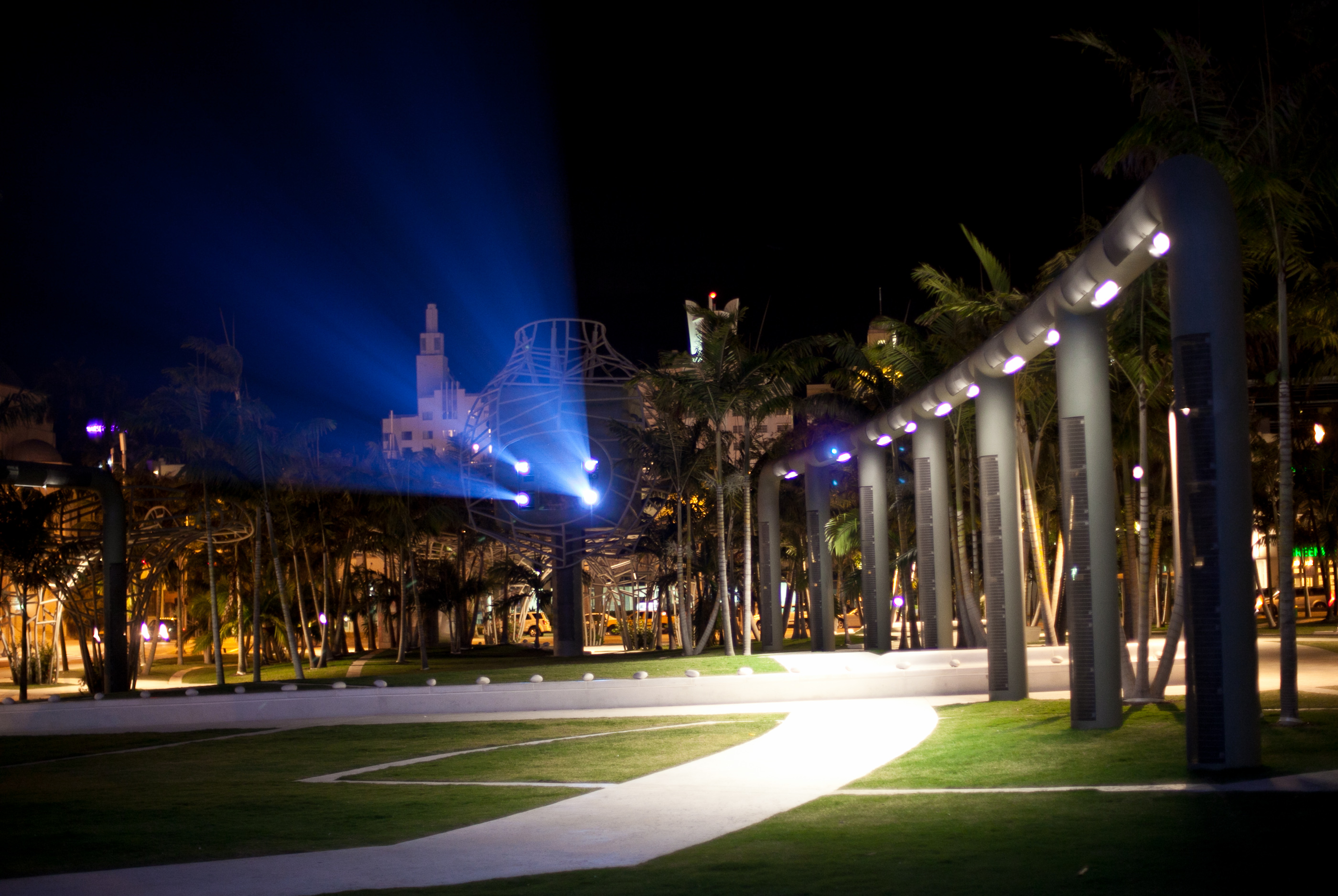 Nighttime exterior of a concert hall with illuminated curved archways, palm trees, and blue light projections.