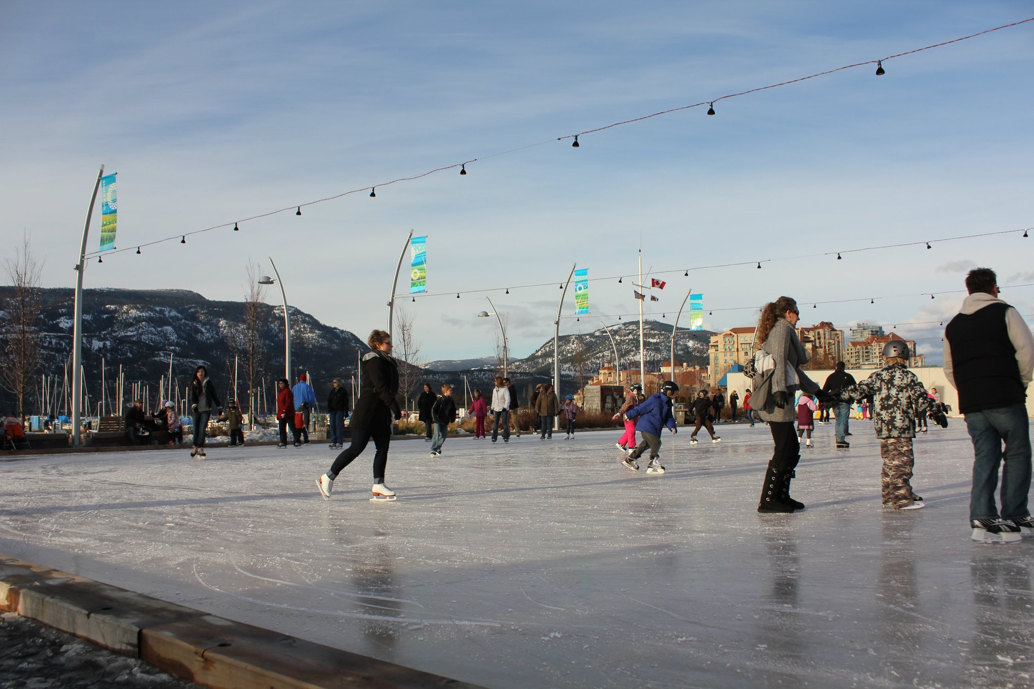 Skaters of various ages glide on an outdoor ice rink with string lights overhead.