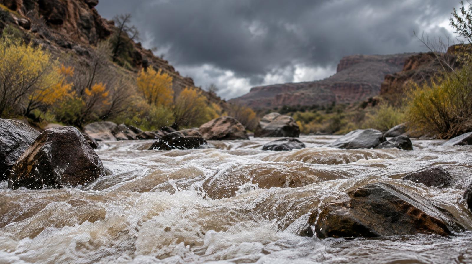 A rushing creek flows through a rocky canyon under dark overcast storm clouds.