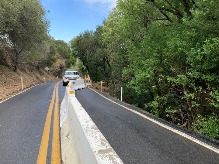 Two-lane road narrowed by white concrete K-rail barriers and orange construction barricades, with a white SUV stopped ahead.