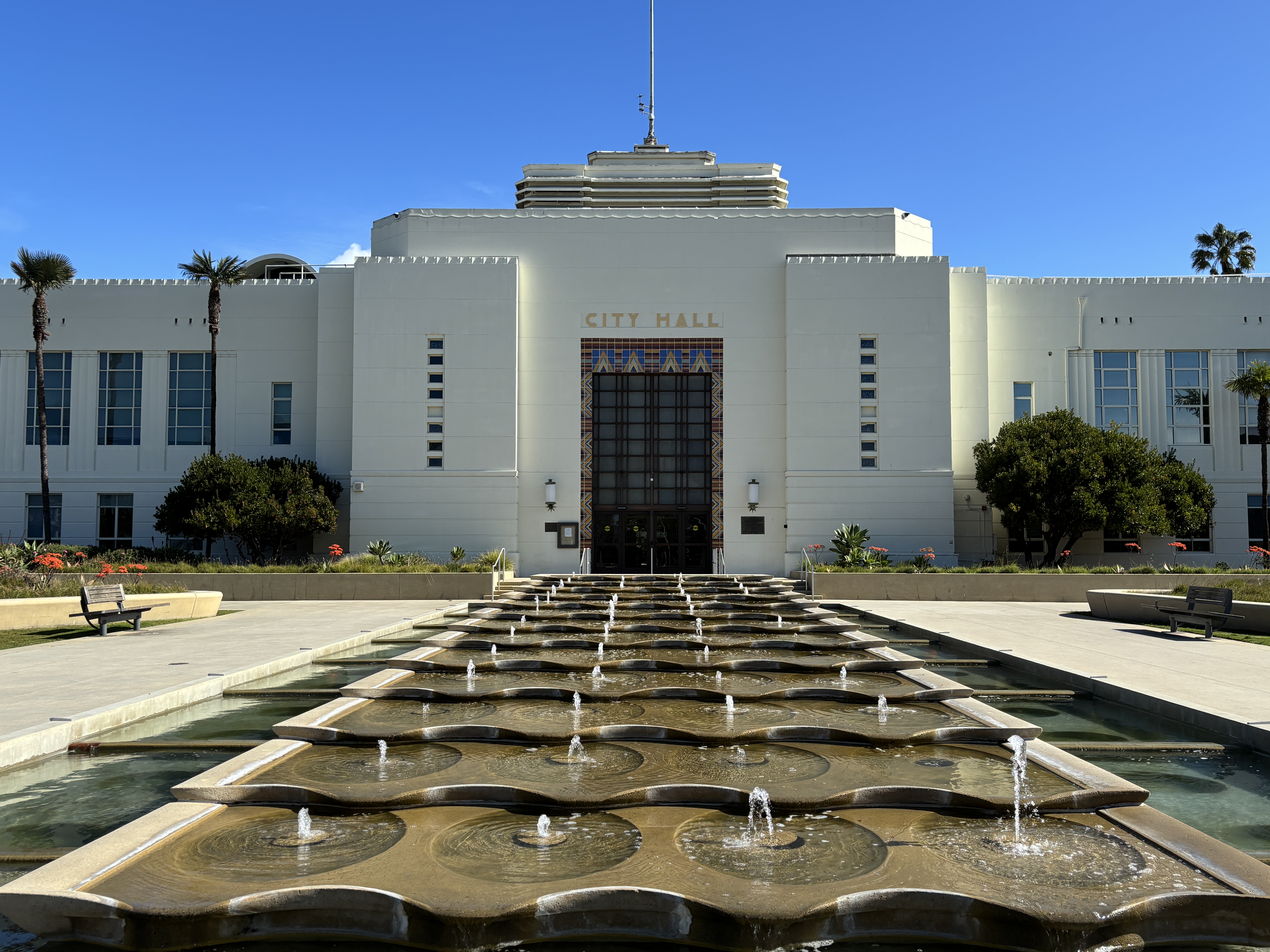 White Art Deco city hall building with fountain and palm trees in front.