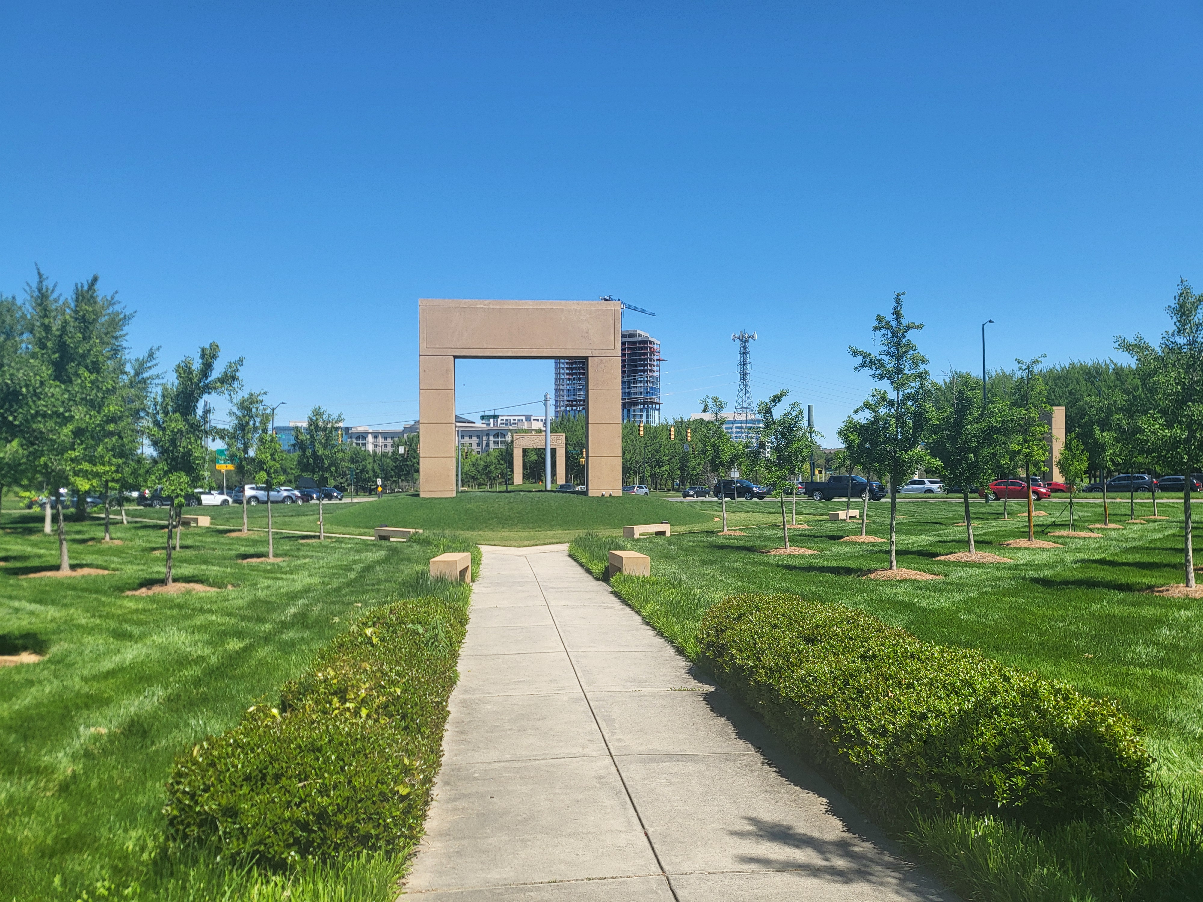 Stone arch monument at the end of a straight walkway flanked by young trees.