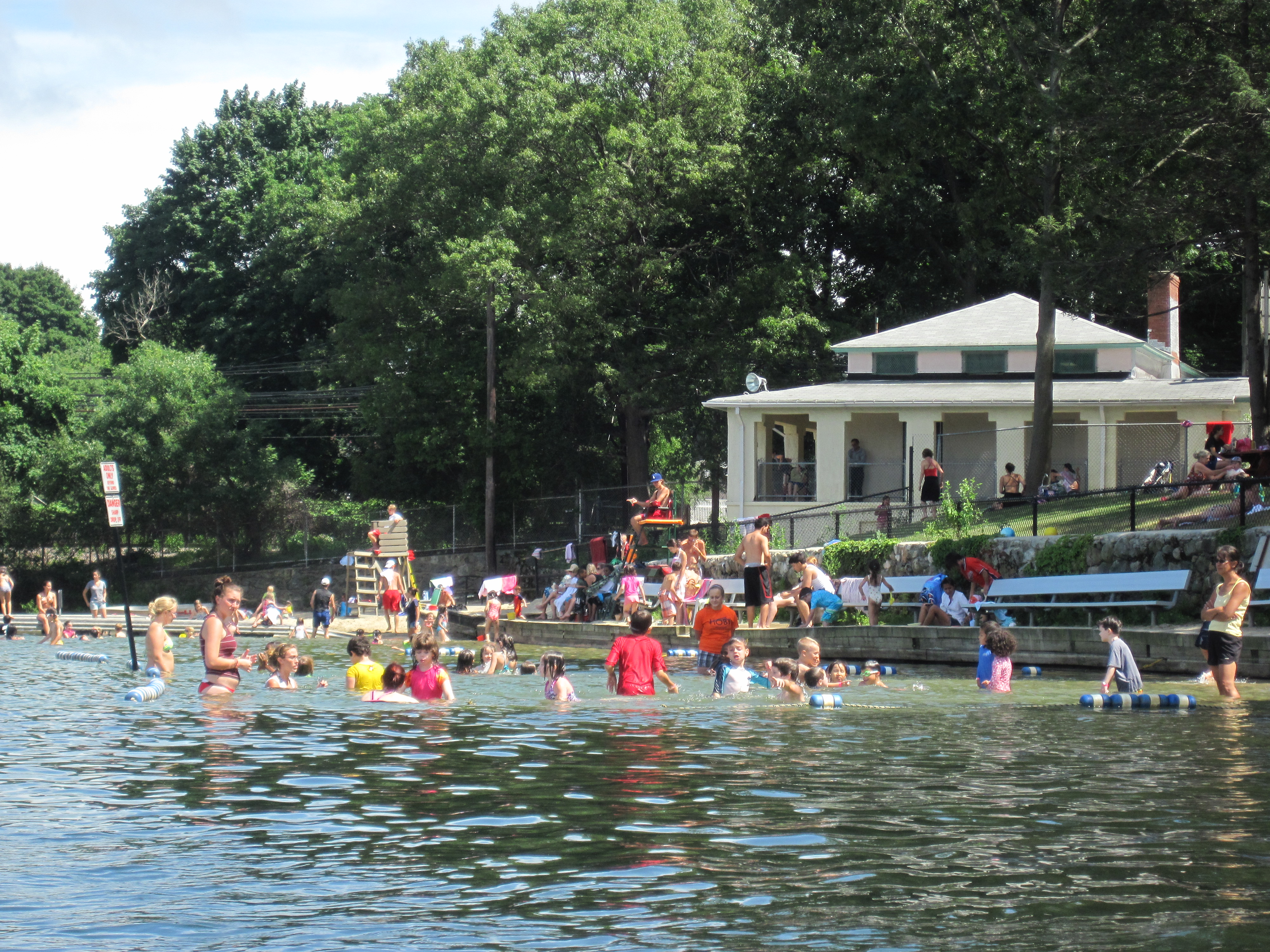 Swimmers at a freshwater lake beach with a white bathhouse building visible among trees.