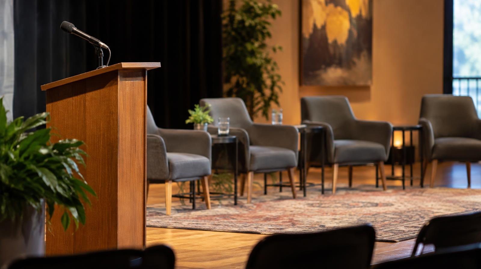 Empty chairs arranged in a semicircle around a moderator's podium in a warmly lit venue.