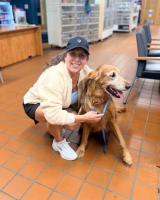 A smiling woman crouches next to a red golden retriever on a blue leash inside an animal shelter.