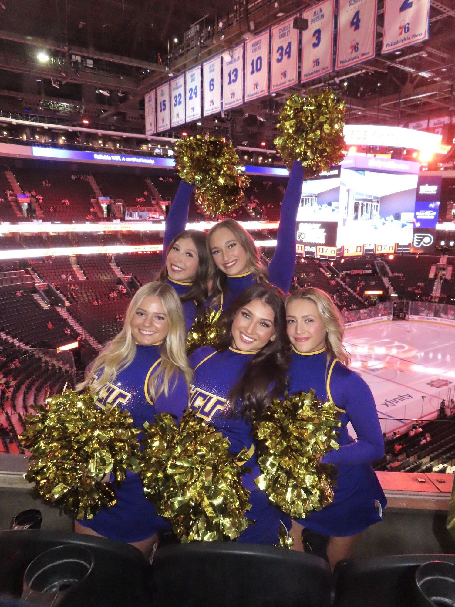 Five women in blue and gold uniforms hold gold pom-poms inside a large sports arena with an ice rink below.