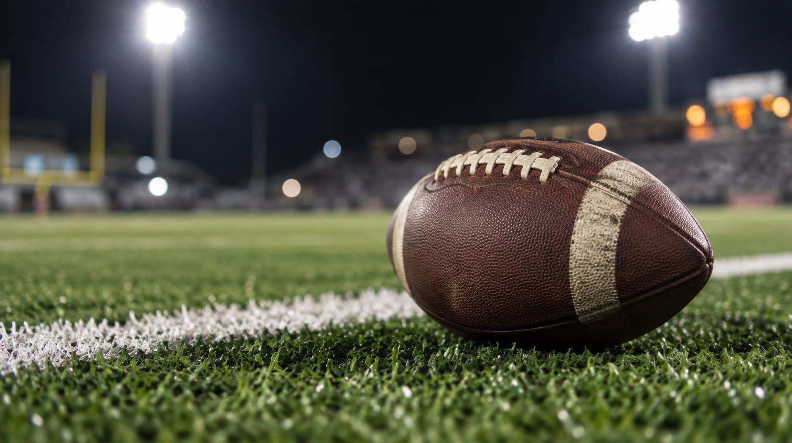 An American football on green artificial turf beside a white yard line under bright stadium lights.